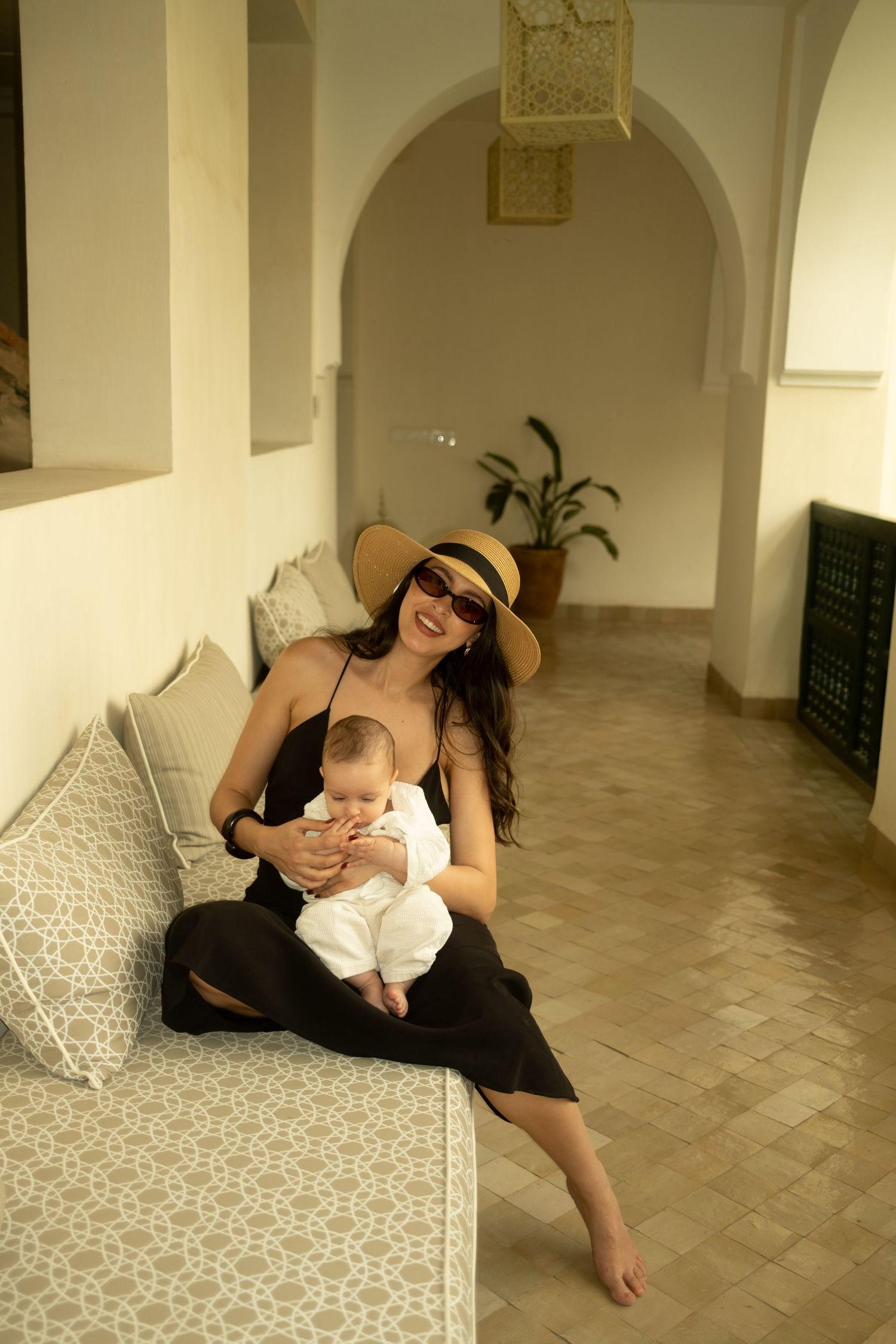 Woman in black dress and sun hat holding a baby on a couch in a Moroccan riad interior.