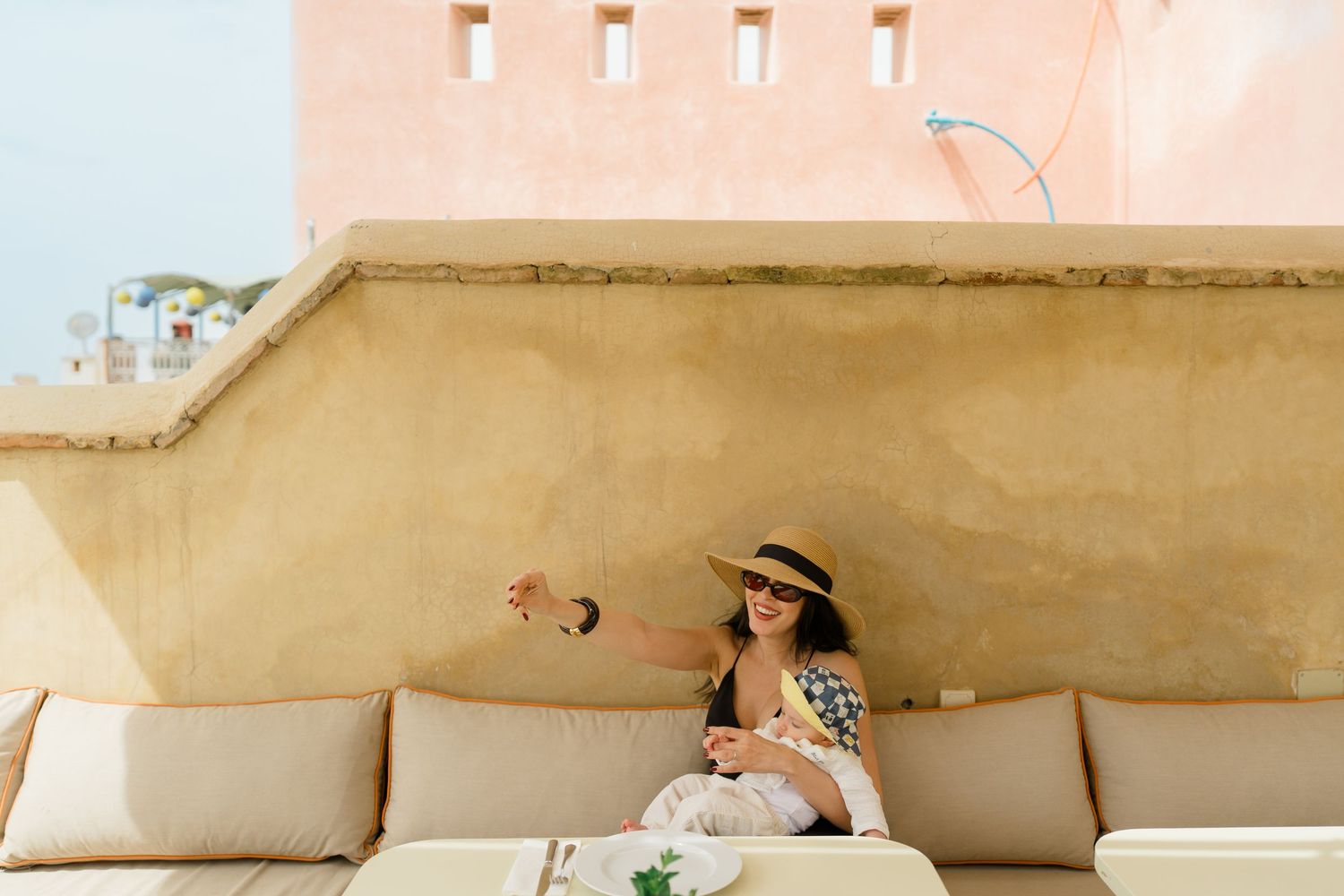 Woman in straw hat and sunglasses relaxing on outdoor terrace sofa, enjoying drinks with Mediterranean architecture backdrop.