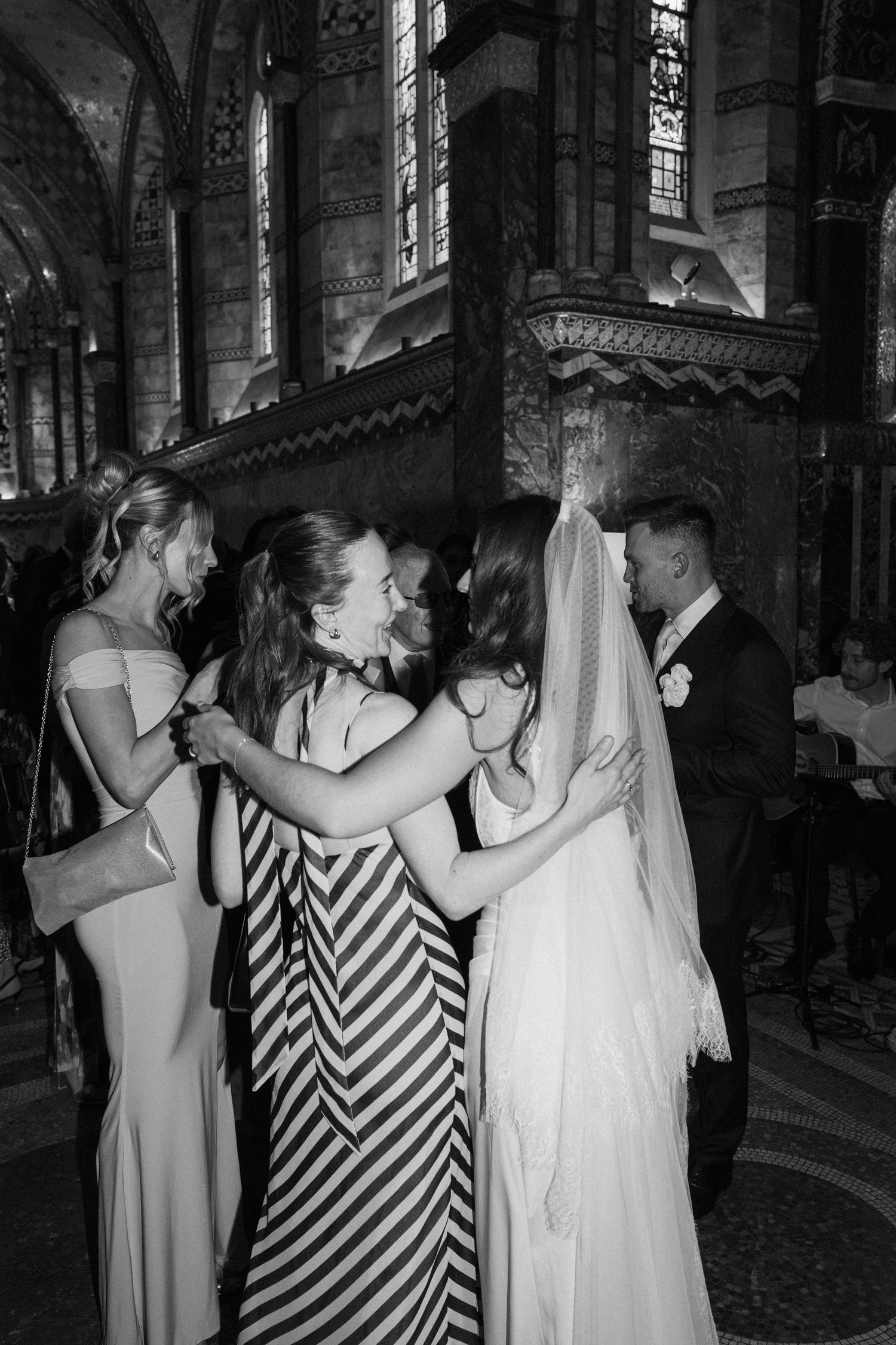 Black and white photo of a bride in white gown embracing wedding guests inside an ornate church.