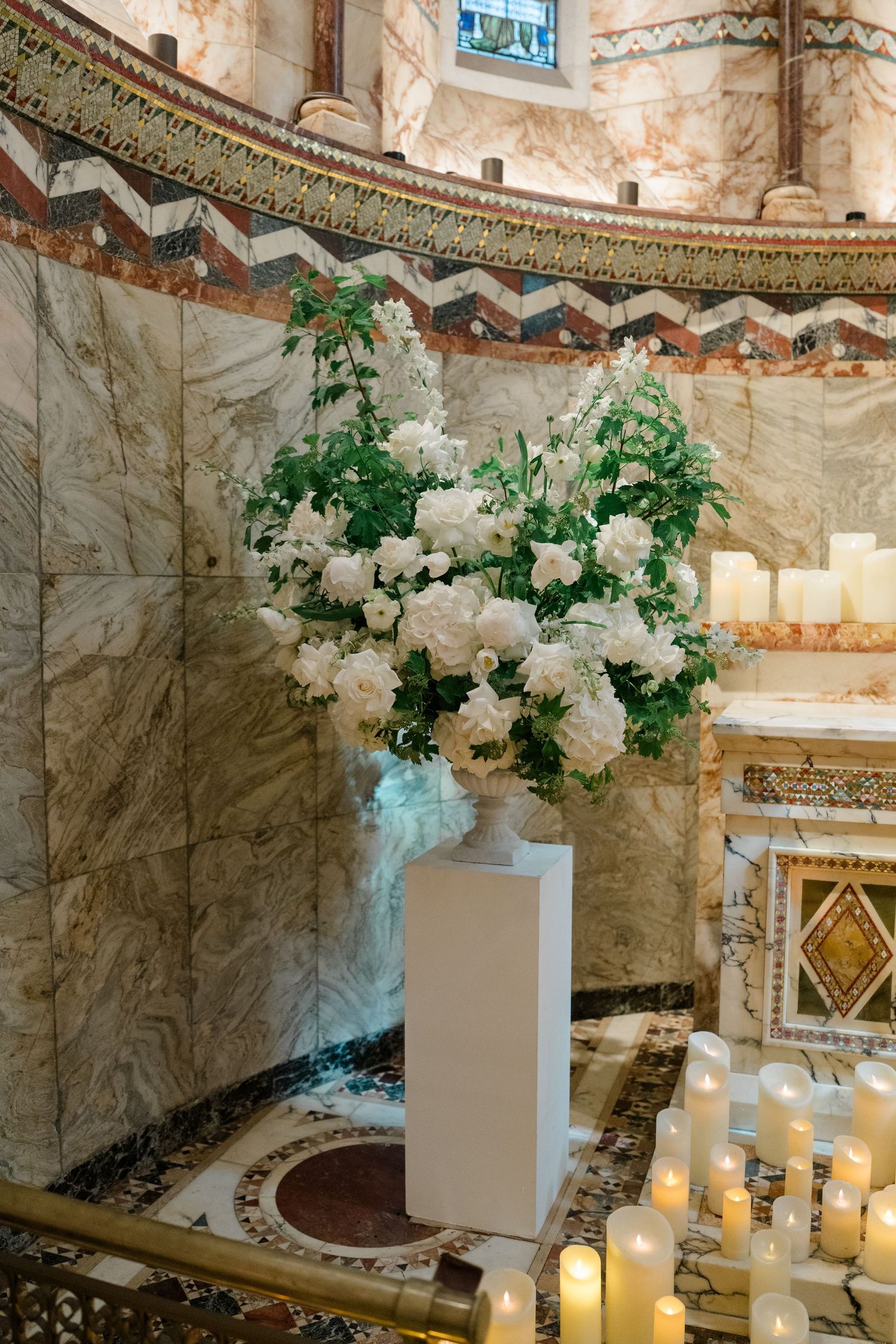 Elegant white floral arrangement with greenery on a white pedestal surrounded by candles in a marble church.