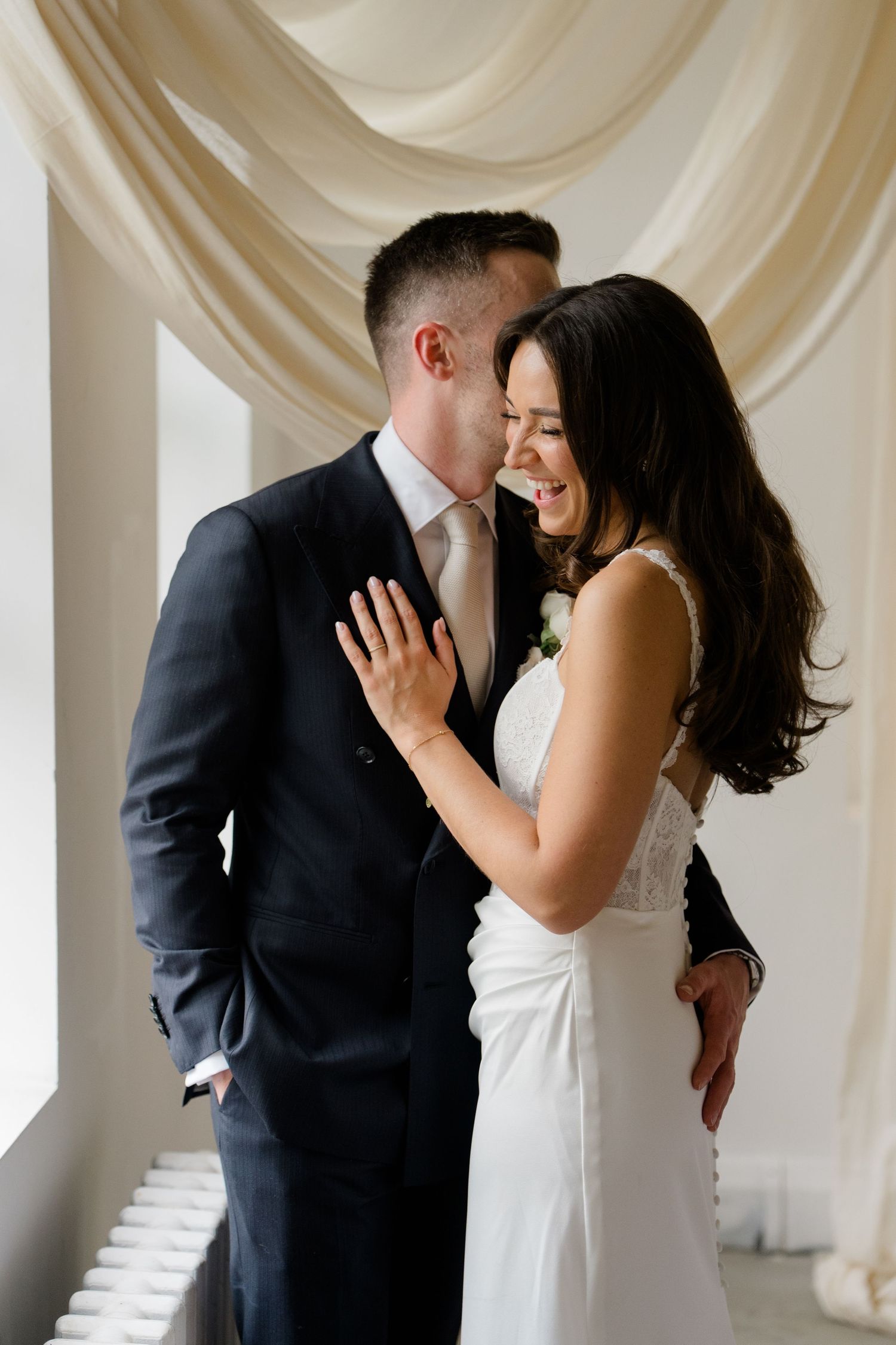 Bride and groom sharing a tender moment, laughing together near draped cream curtains.