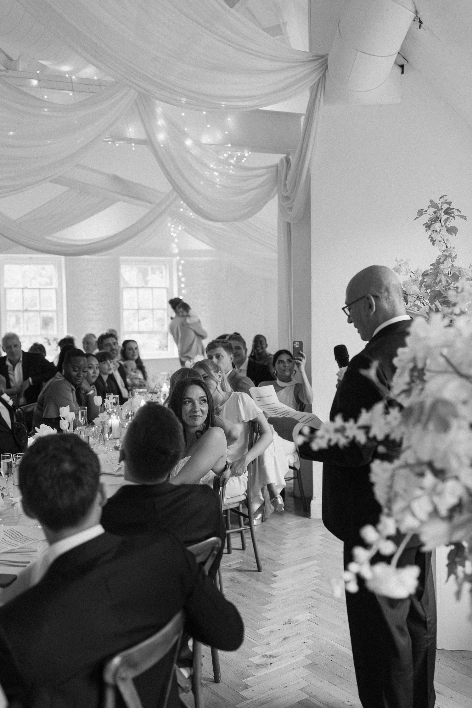 Father of bride giving speech to seated wedding guests in draped reception hall with fairy lights.