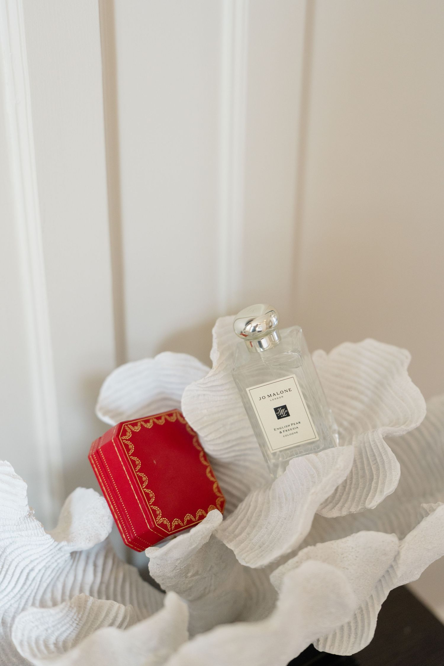 Jo Malone perfume bottle and red Cartier ring box on white fabric background.