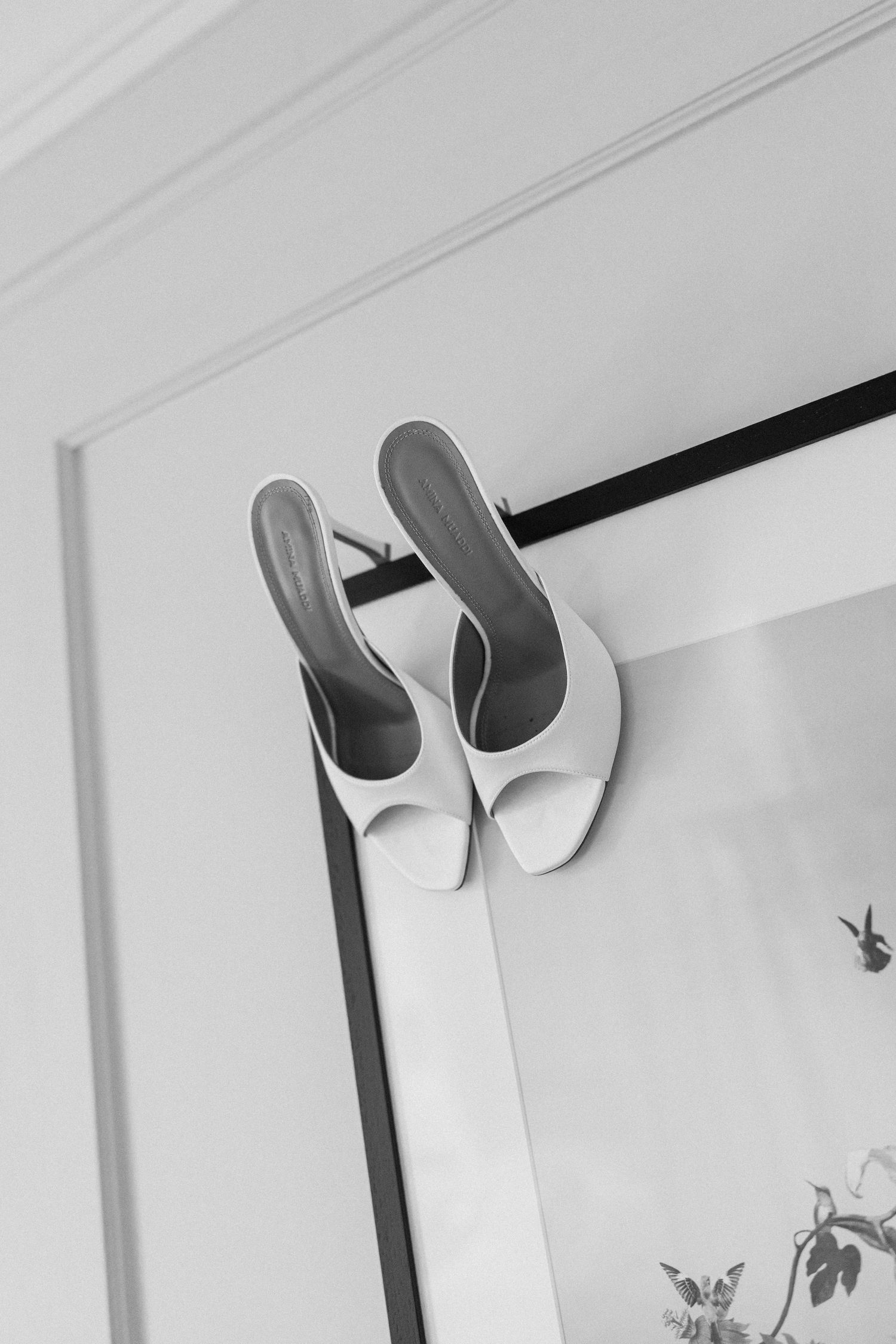 White open-toe mule heels hanging on a mirror frame, black and white minimalist photography.