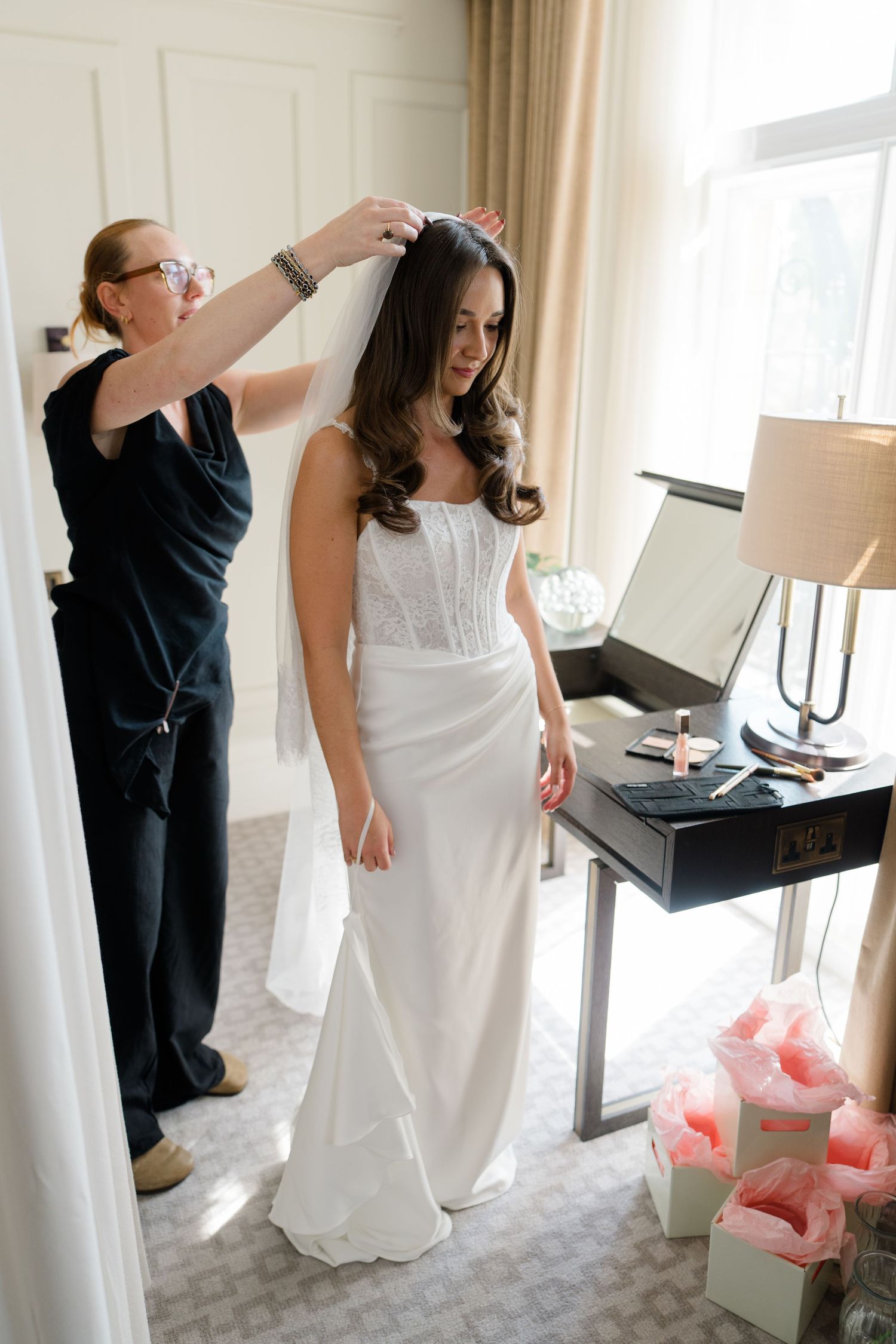 A stylist places a veil on a bride in a sleek white wedding gown in a hotel suite.