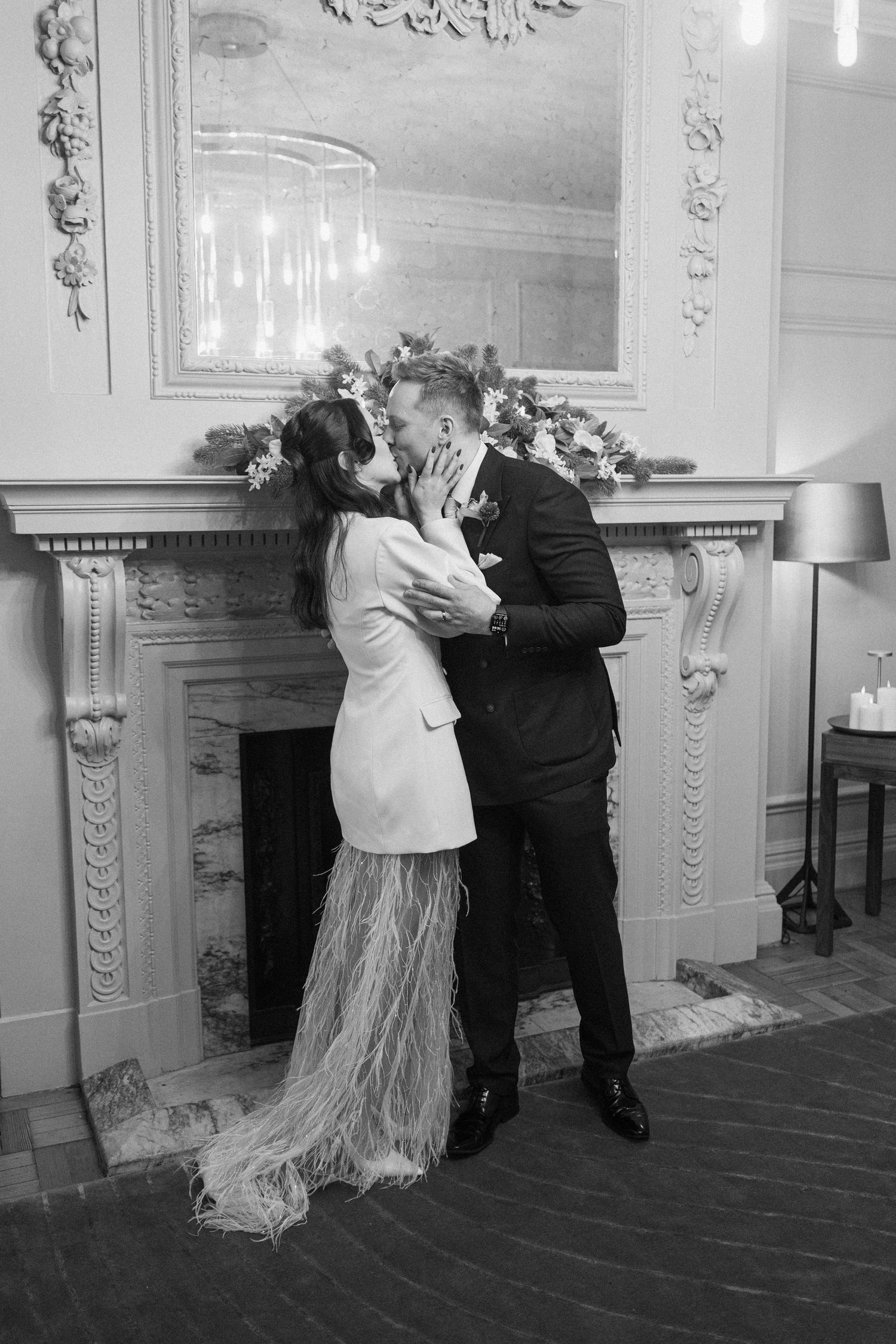 Bride and groom share a kiss in front of an ornate fireplace, bride wearing a white blazer dress with feather hem.