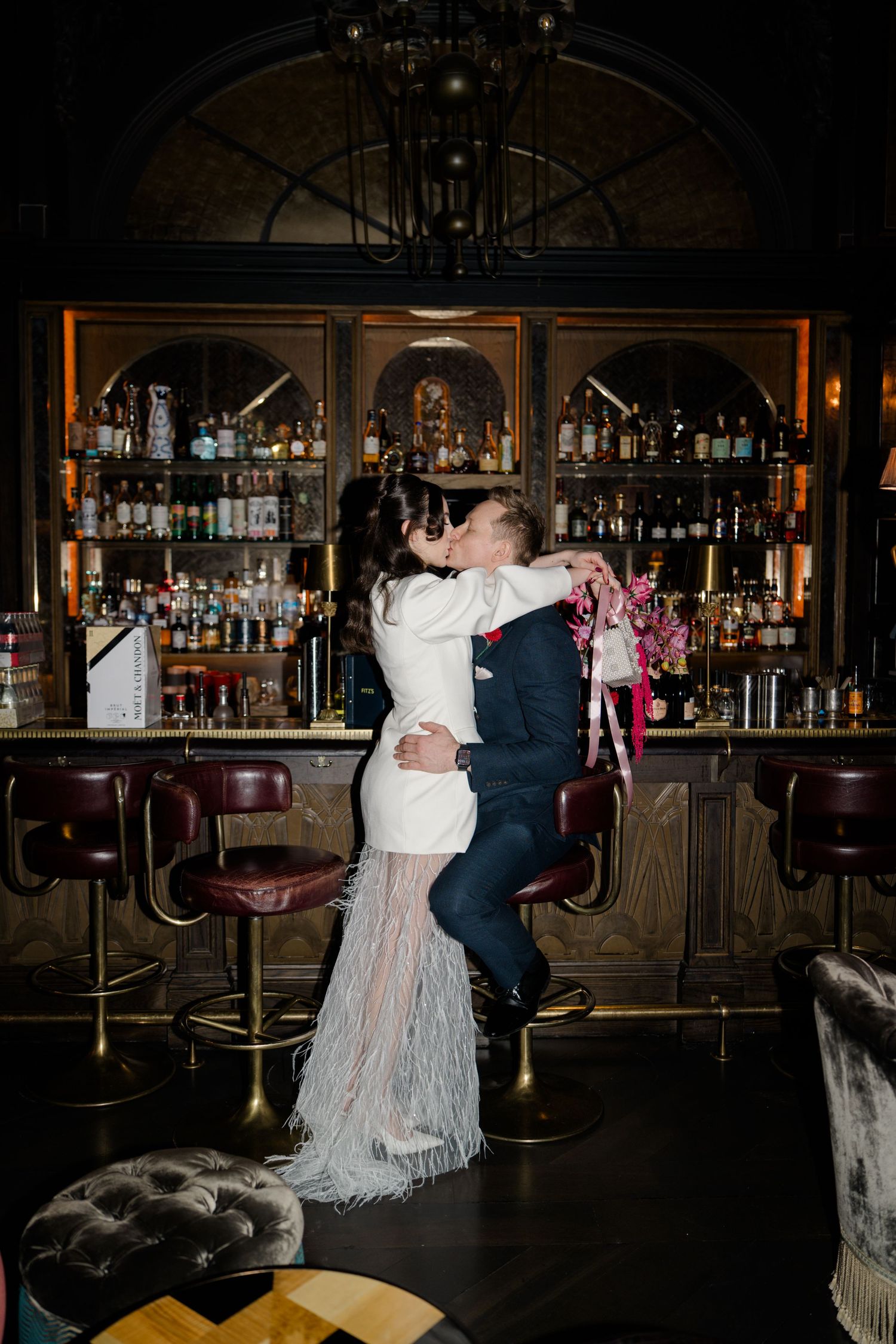Bride and groom share a romantic kiss at a dark, moody cocktail bar on their wedding day.