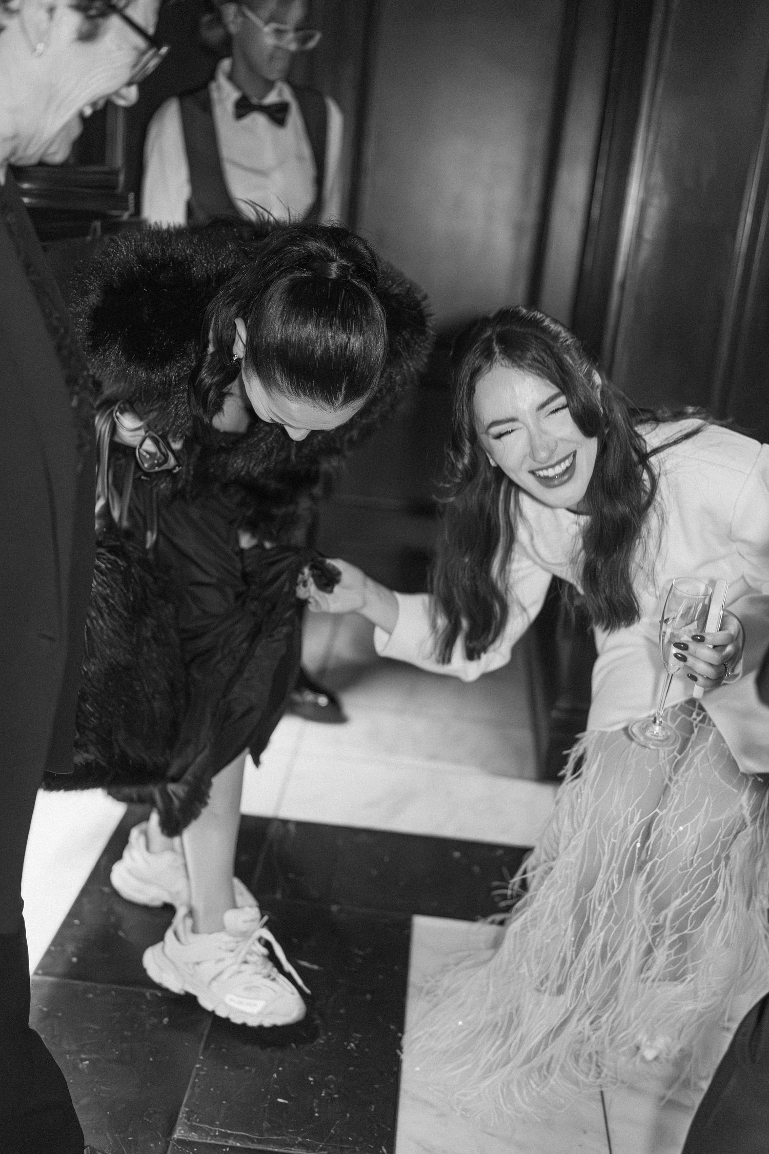 Black and white photo of laughing wedding guests in formal attire gathered together at elegant event.