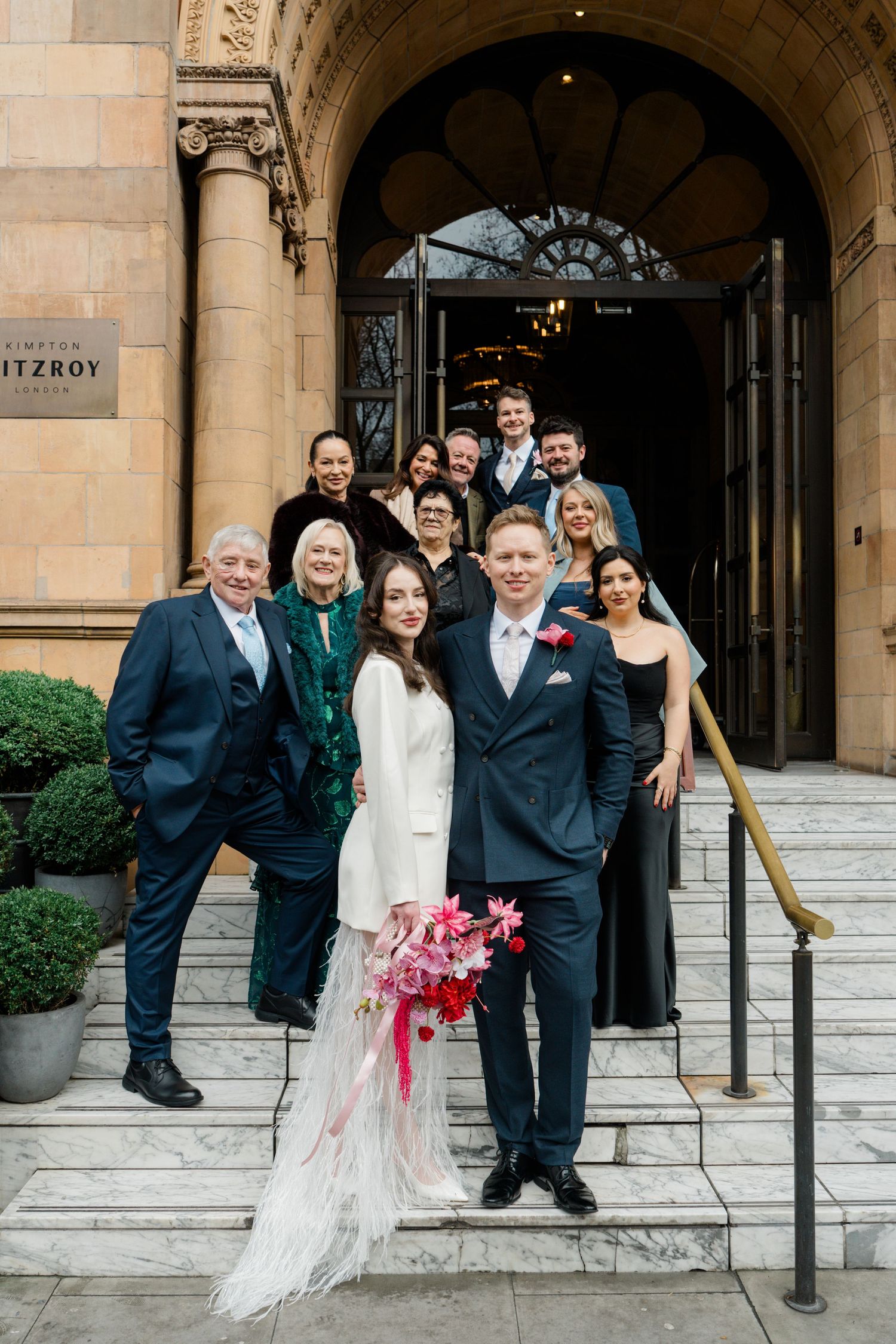 Wedding party poses on steps outside The Fitzroy London venue with bride in white feather dress and groom in navy suit.