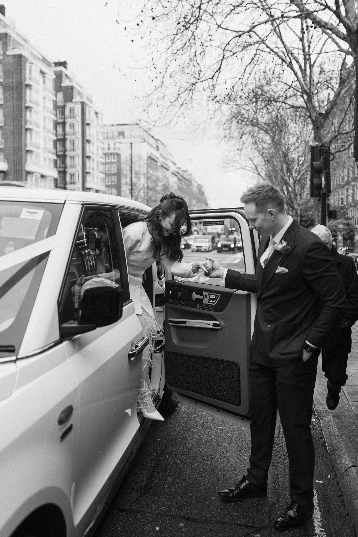 Black and white photo of groom helping bride exit a white taxi cab on a city street.