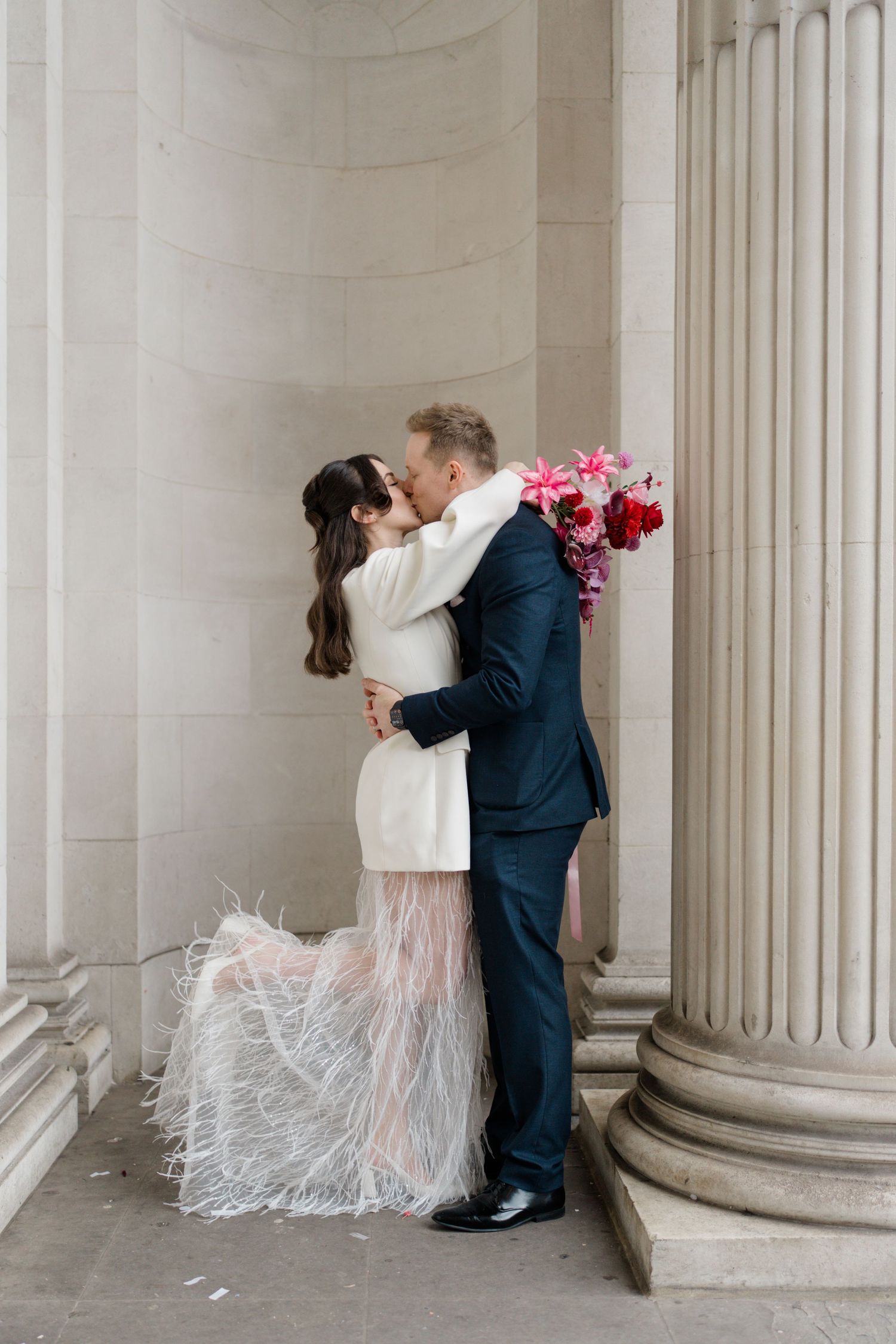 Bride and groom kissing between stone columns, bride wearing white blazer with feathered skirt.