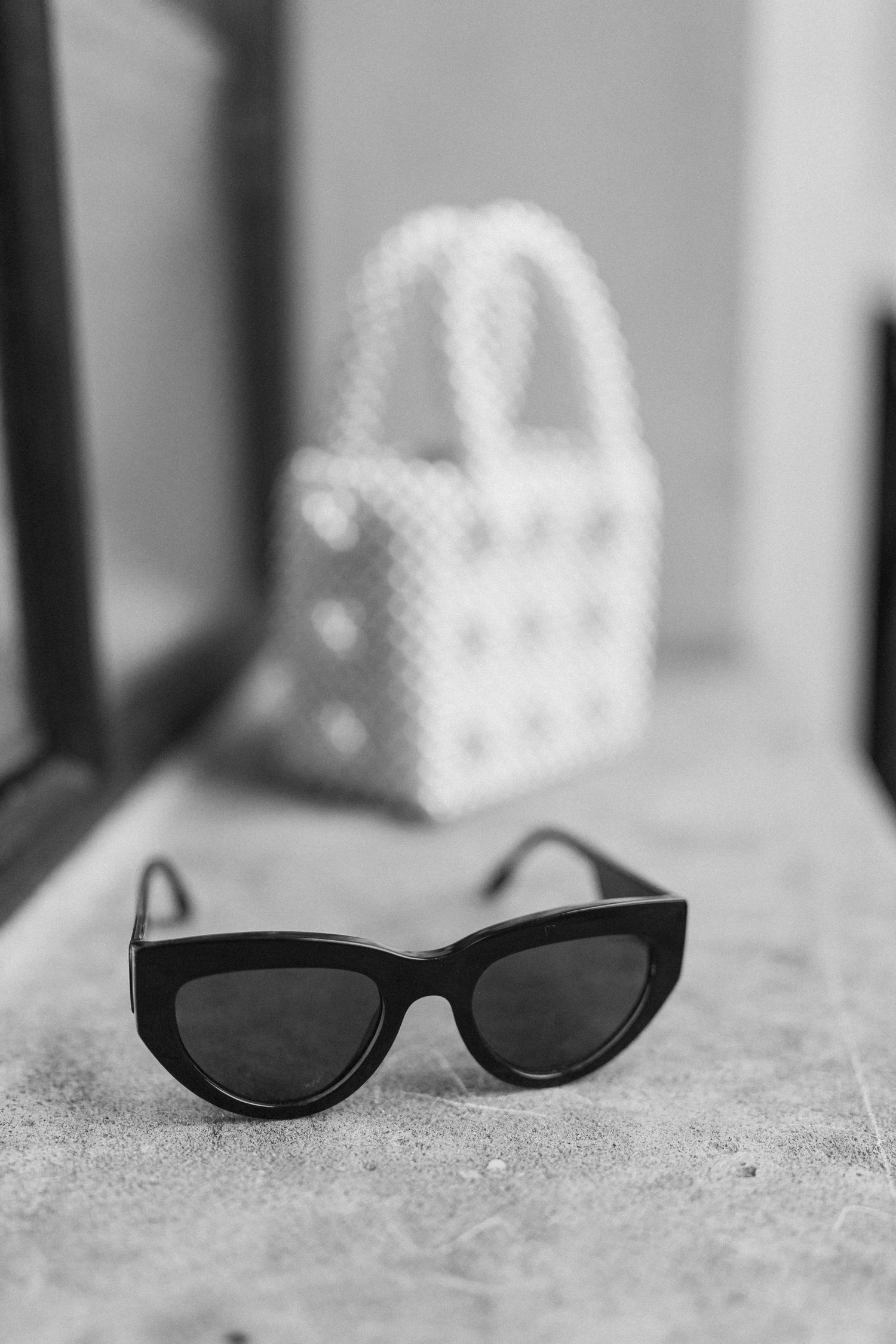 Black and white photo of cat-eye sunglasses with pearl handbag in background.