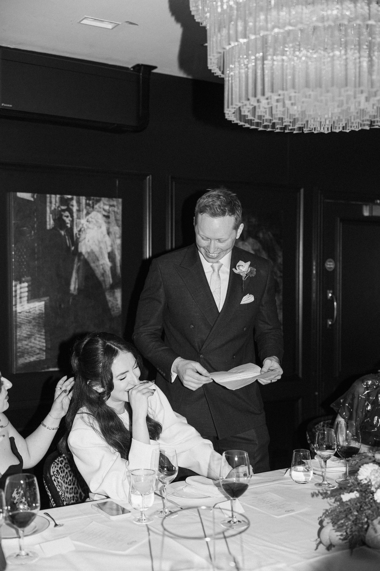 Black and white photo of groom reading heartfelt speech to bride at intimate wedding dinner.