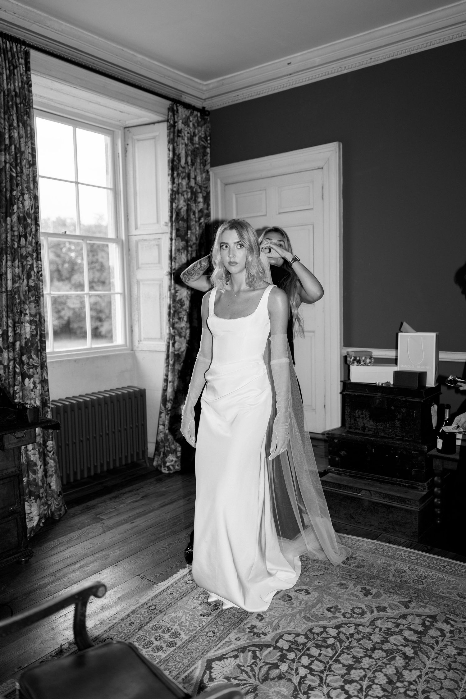 Bride in a sleek white gown being helped by assistant in a classic room, captured in black and white.