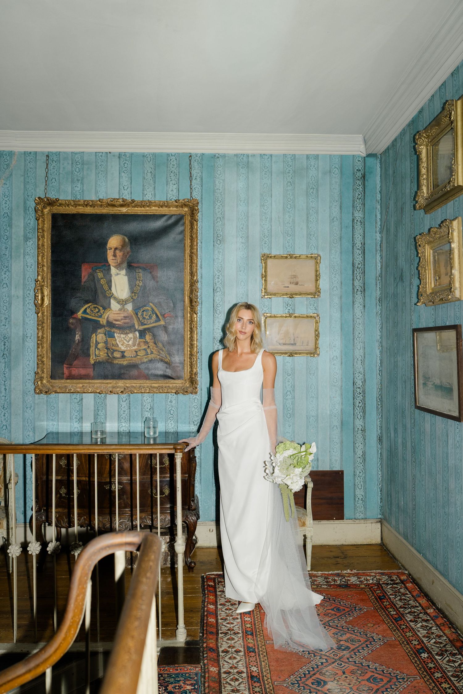 Bride in white gown poses on staircase beside large portrait painting on blue walls.