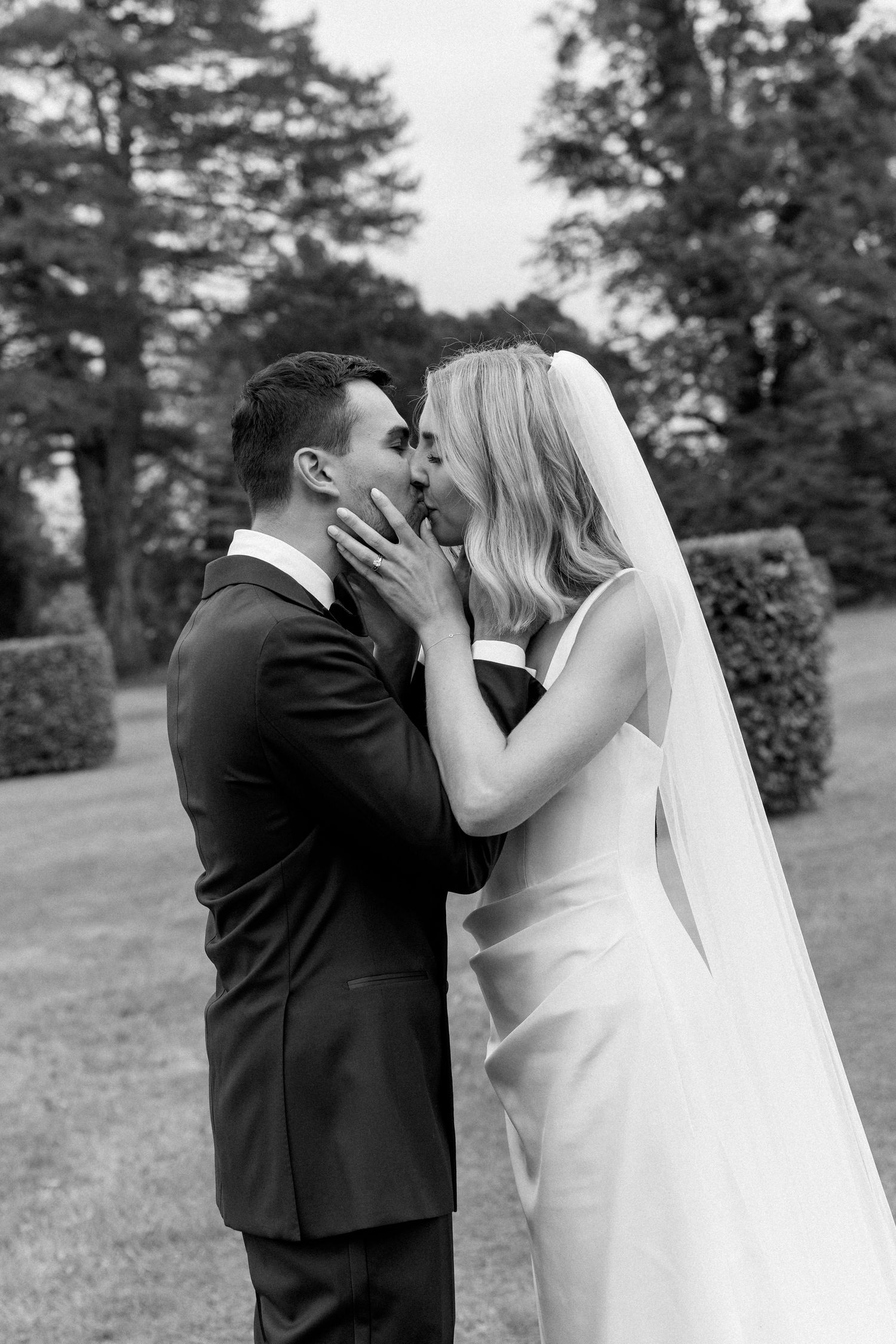 Bride and groom sharing a romantic kiss in a formal garden with manicured hedges.