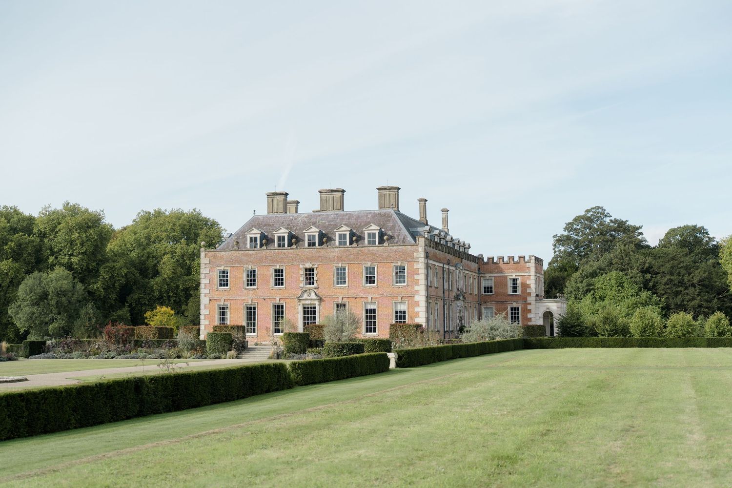 Grand historic English manor house with manicured lawn and hedgerows under a clear sky.