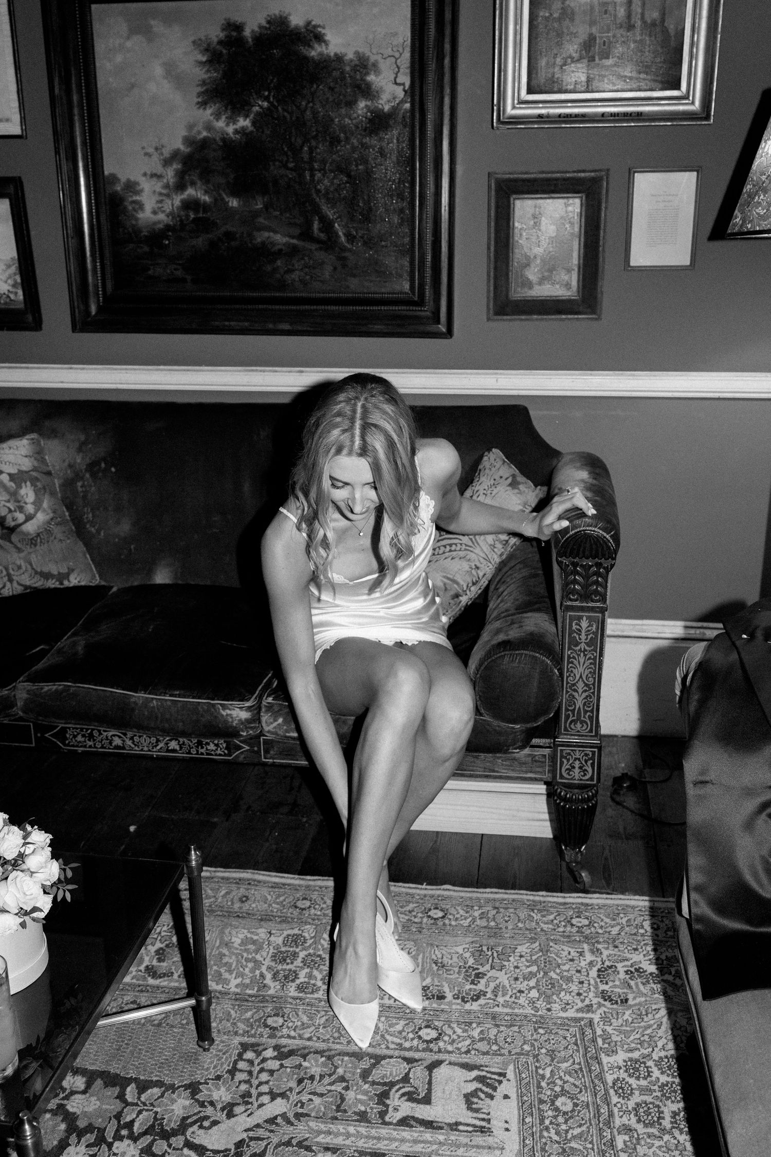 Bride in white dress sits on an ornate sofa smiling while putting on her wedding shoes.