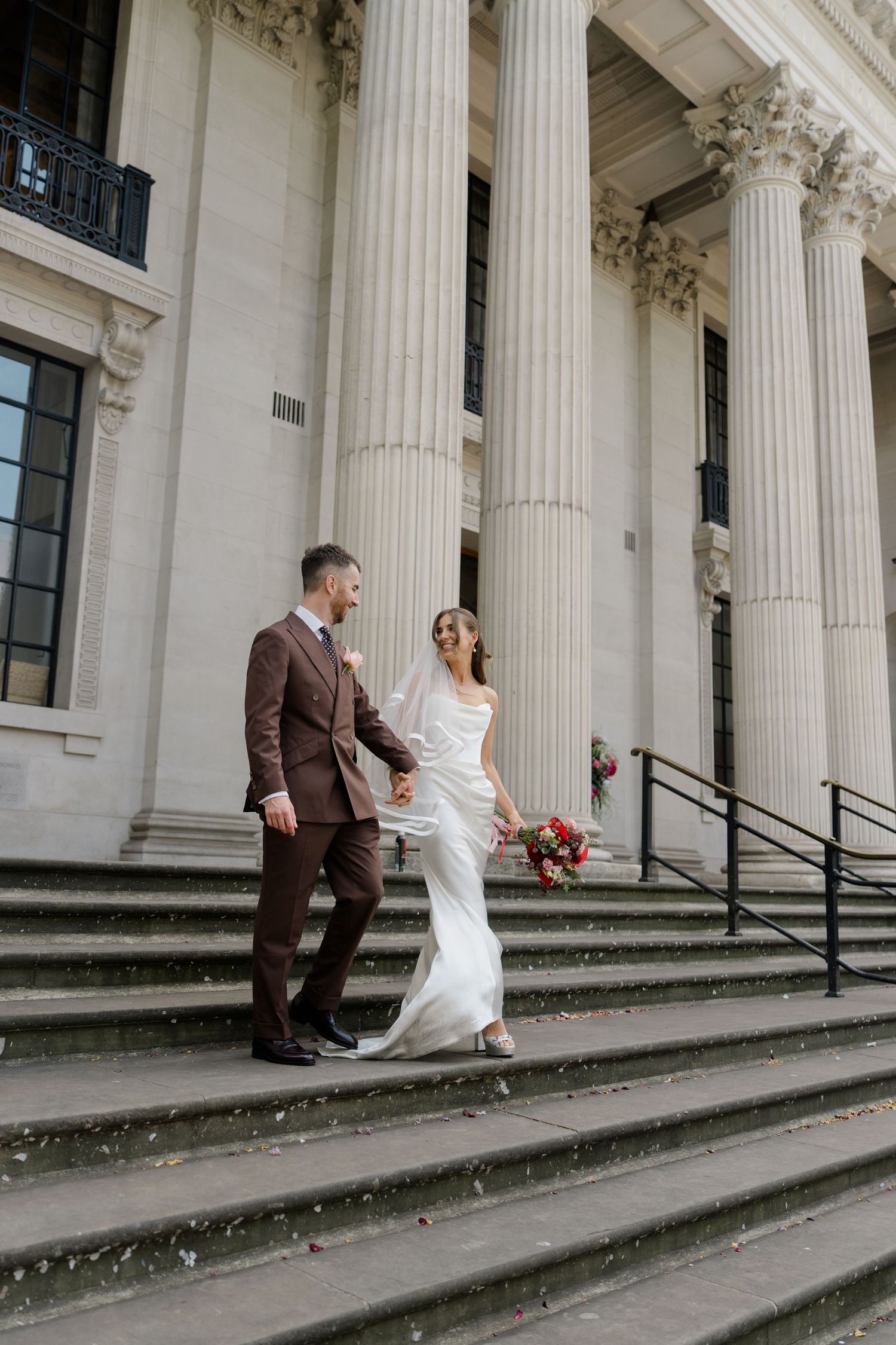Bride and groom holding hands walking down stone steps of a grand neoclassical building with tall columns.