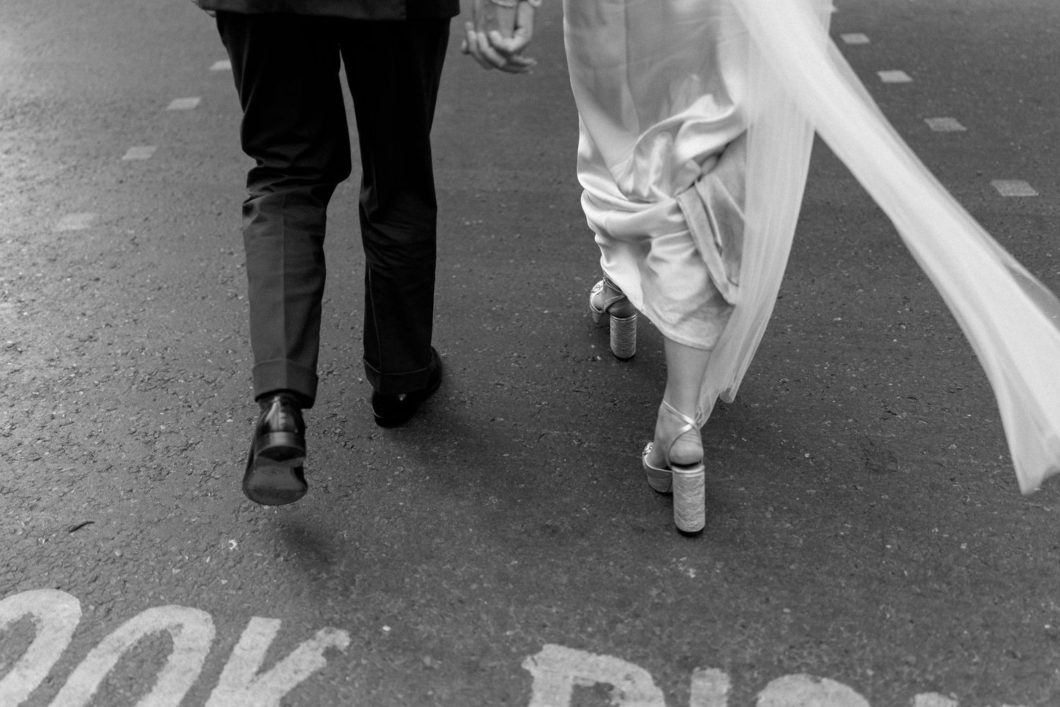 Close-up of bride in platform heels and groom walking across London street crossing in black and white.