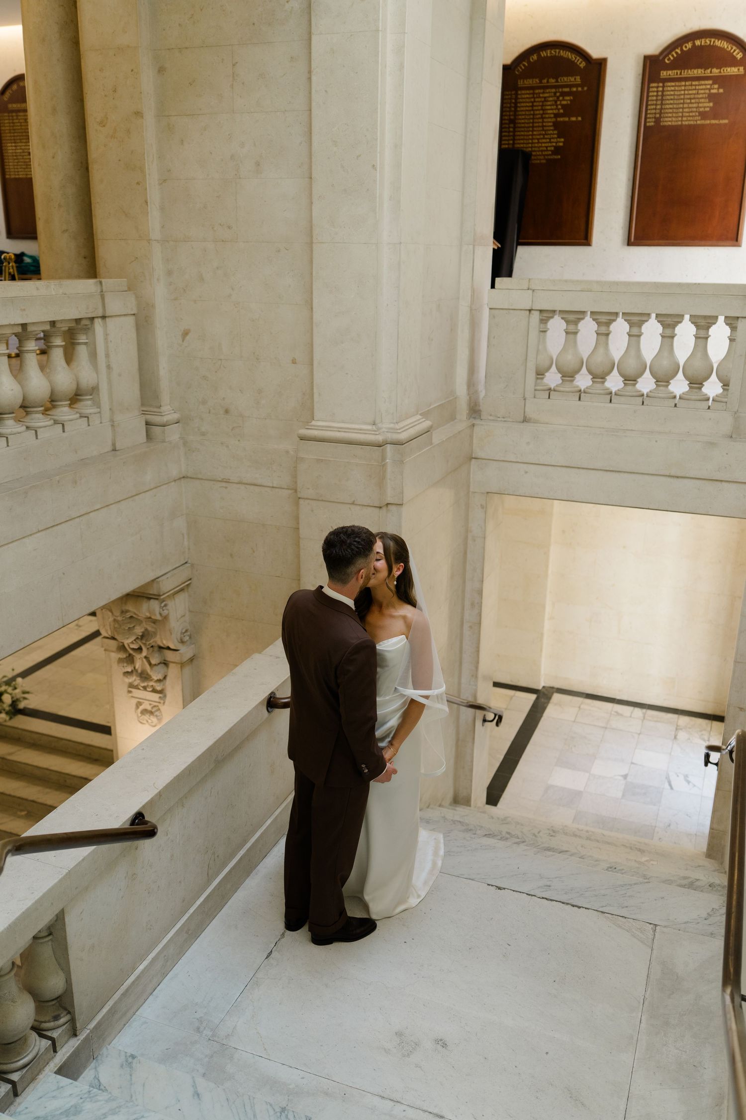 Bride and groom share a romantic kiss on marble stairs in an elegant wedding venue.