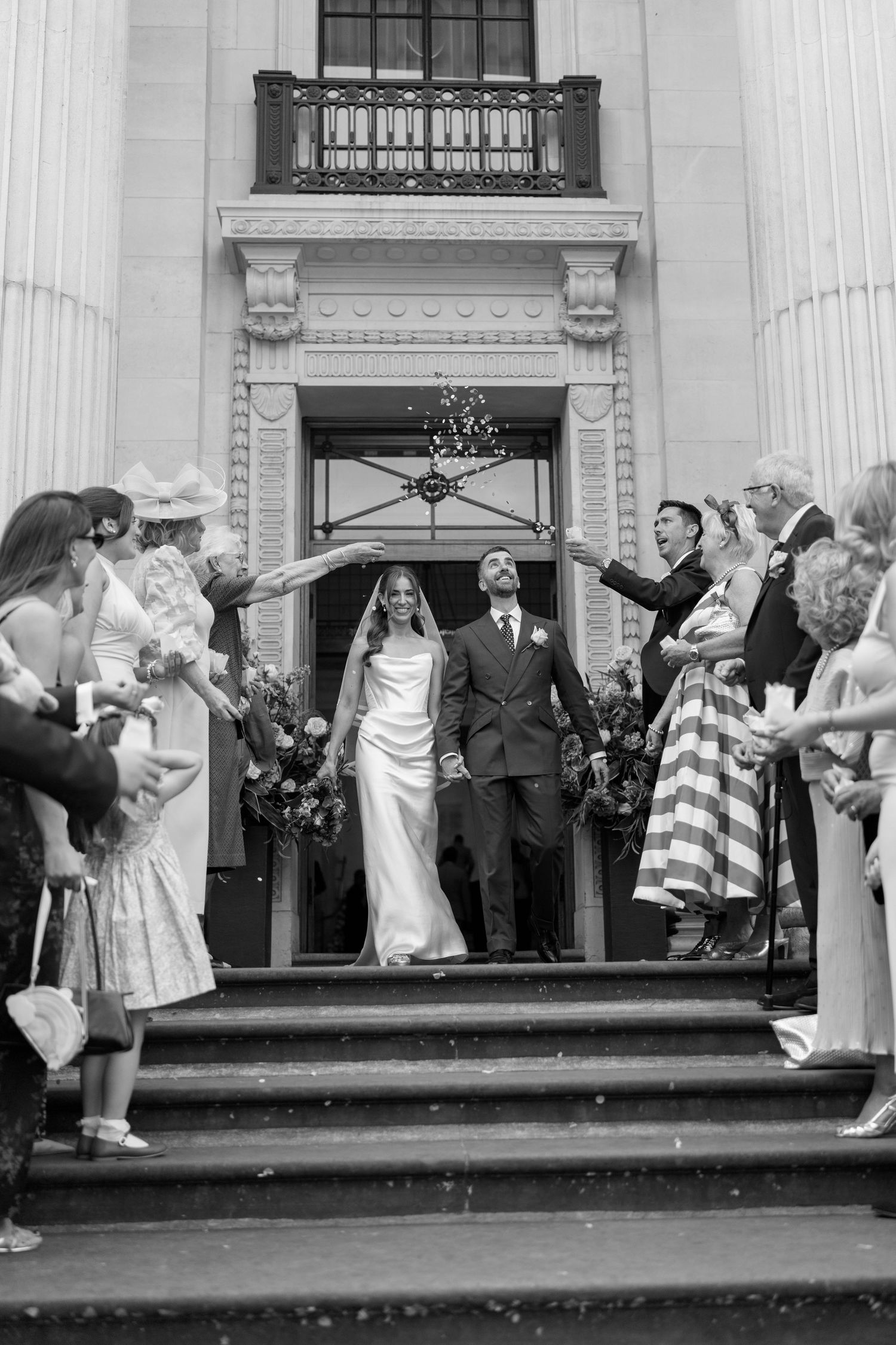 Black and white photo of newlyweds exiting a grand building as guests celebrate with confetti.