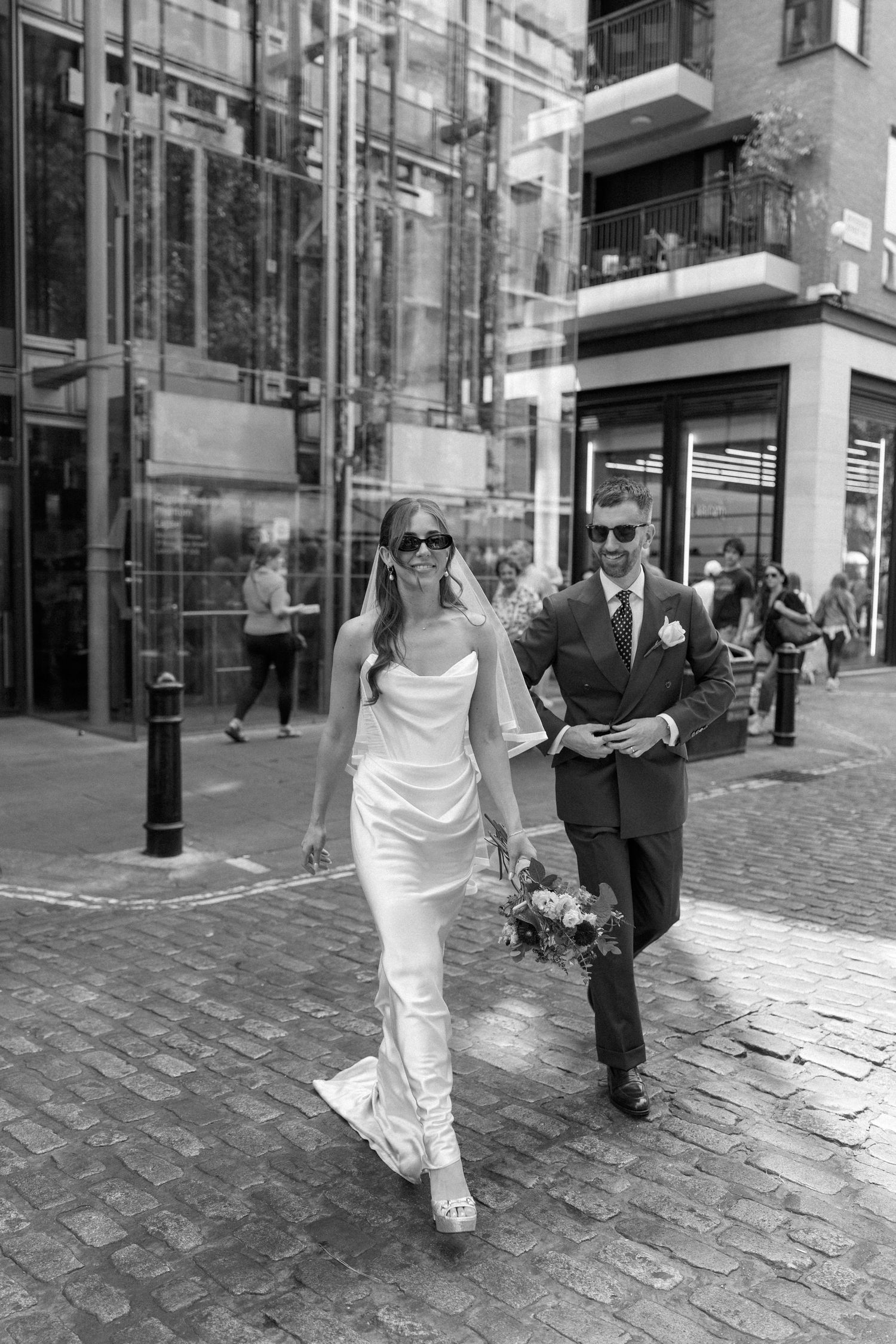 Stylish bride in white gown and sunglasses walks cobblestone street with groom in London city.