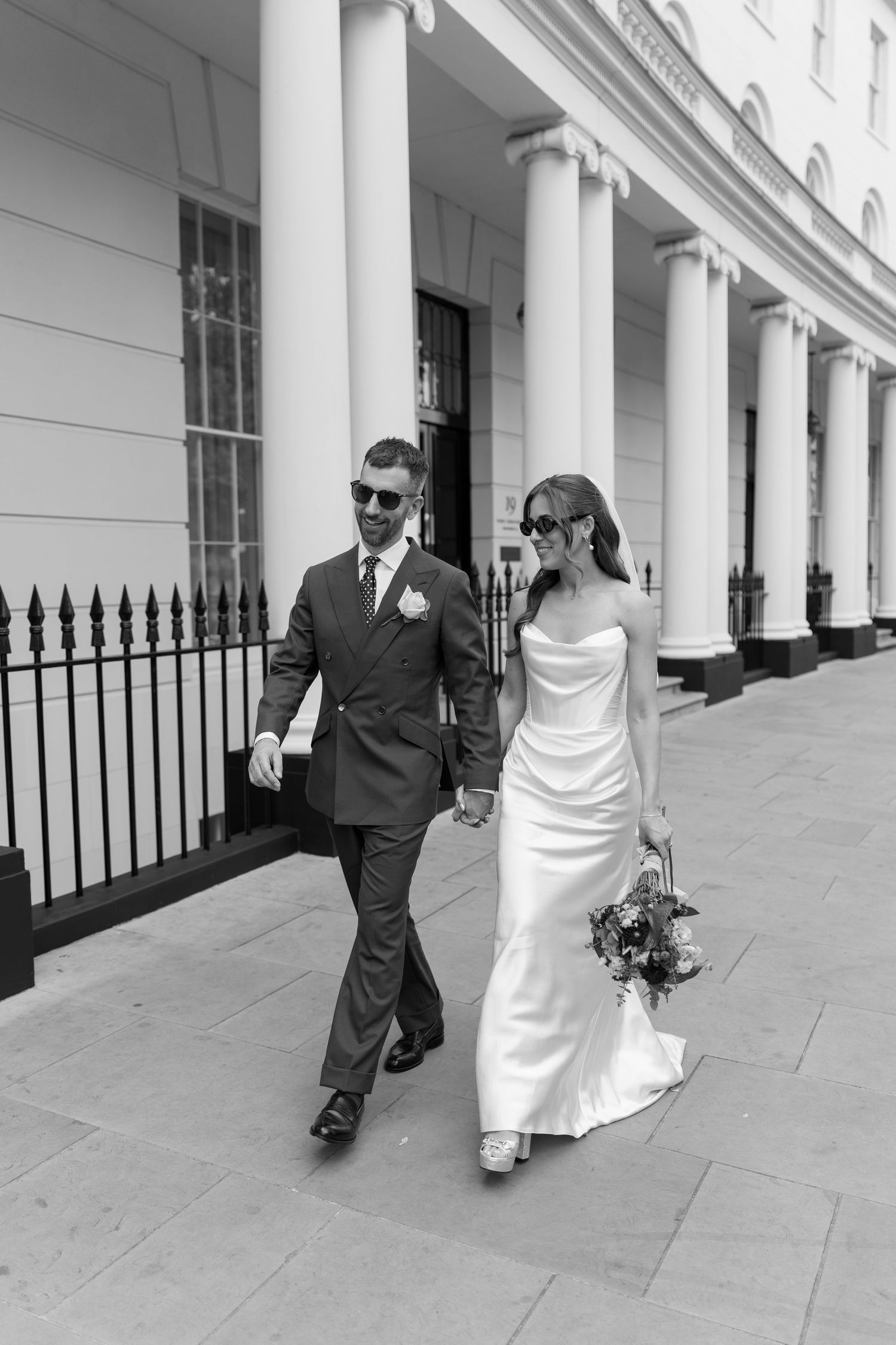 Black and white photo of newlyweds in sunglasses walking hand in hand past elegant white colonnade.