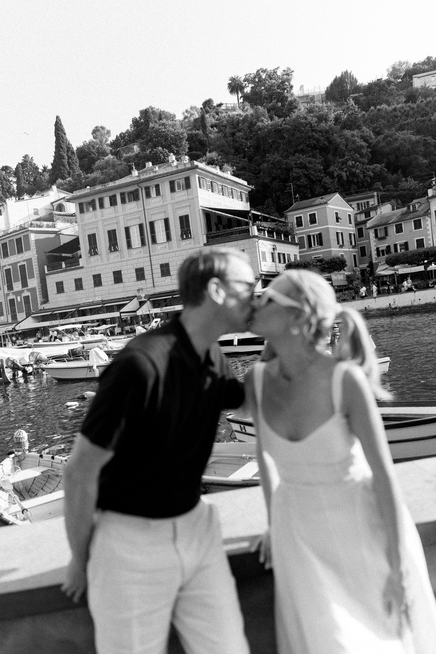 Black and white photo of couple kissing by Portofino harbor, boats and hillside village in background.