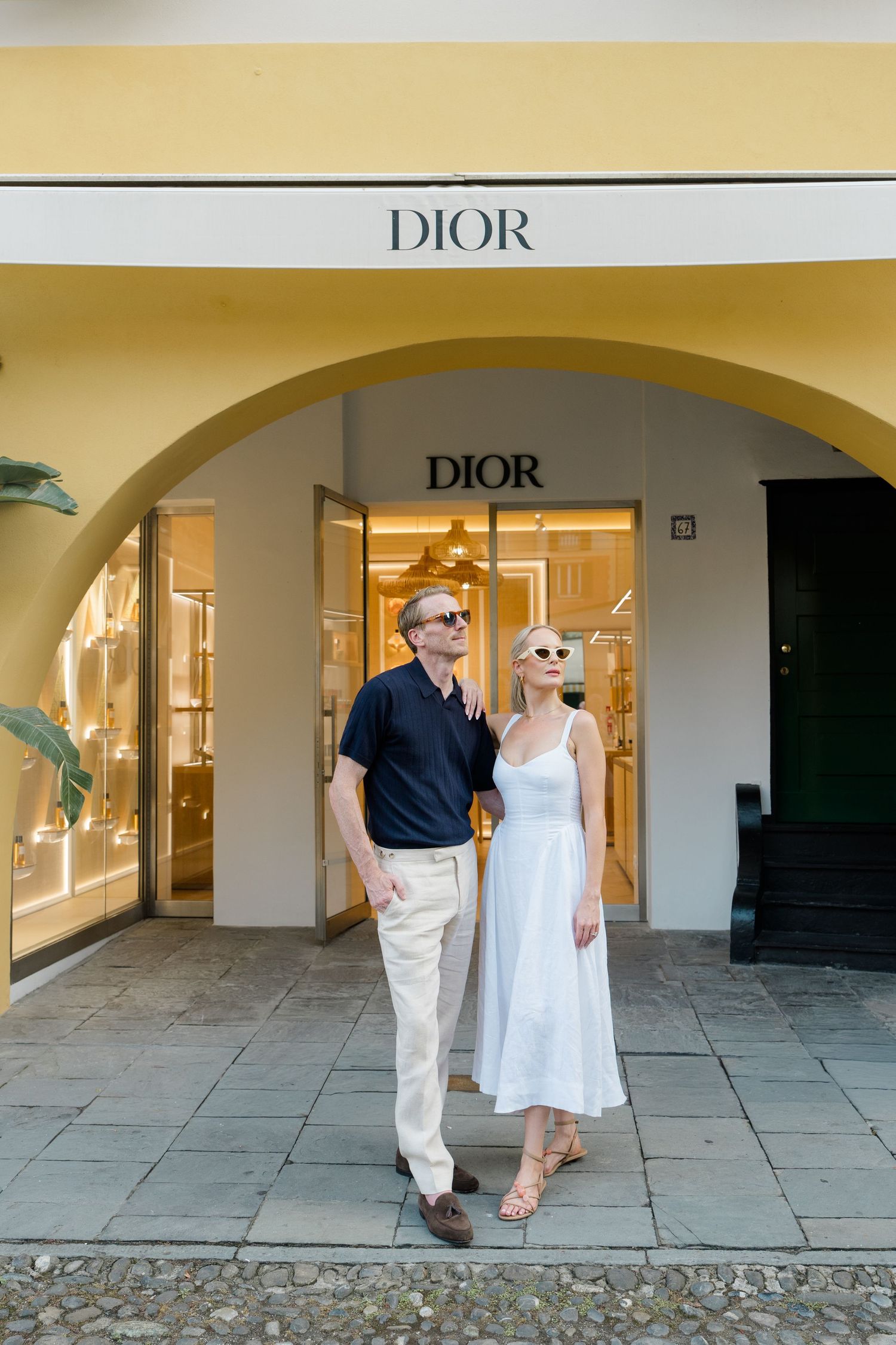 Elegant couple in sunglasses posing under arched entrance of Dior boutique in Portofino, Italy.
