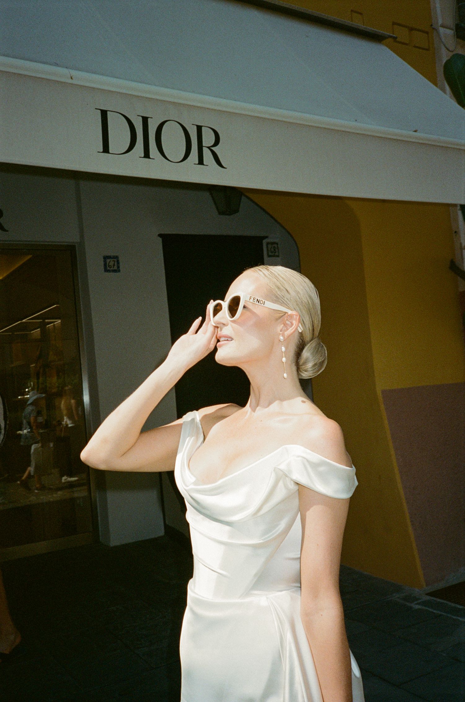 Blonde bride in white satin off-shoulder gown and cat-eye sunglasses posing outside a Dior boutique.