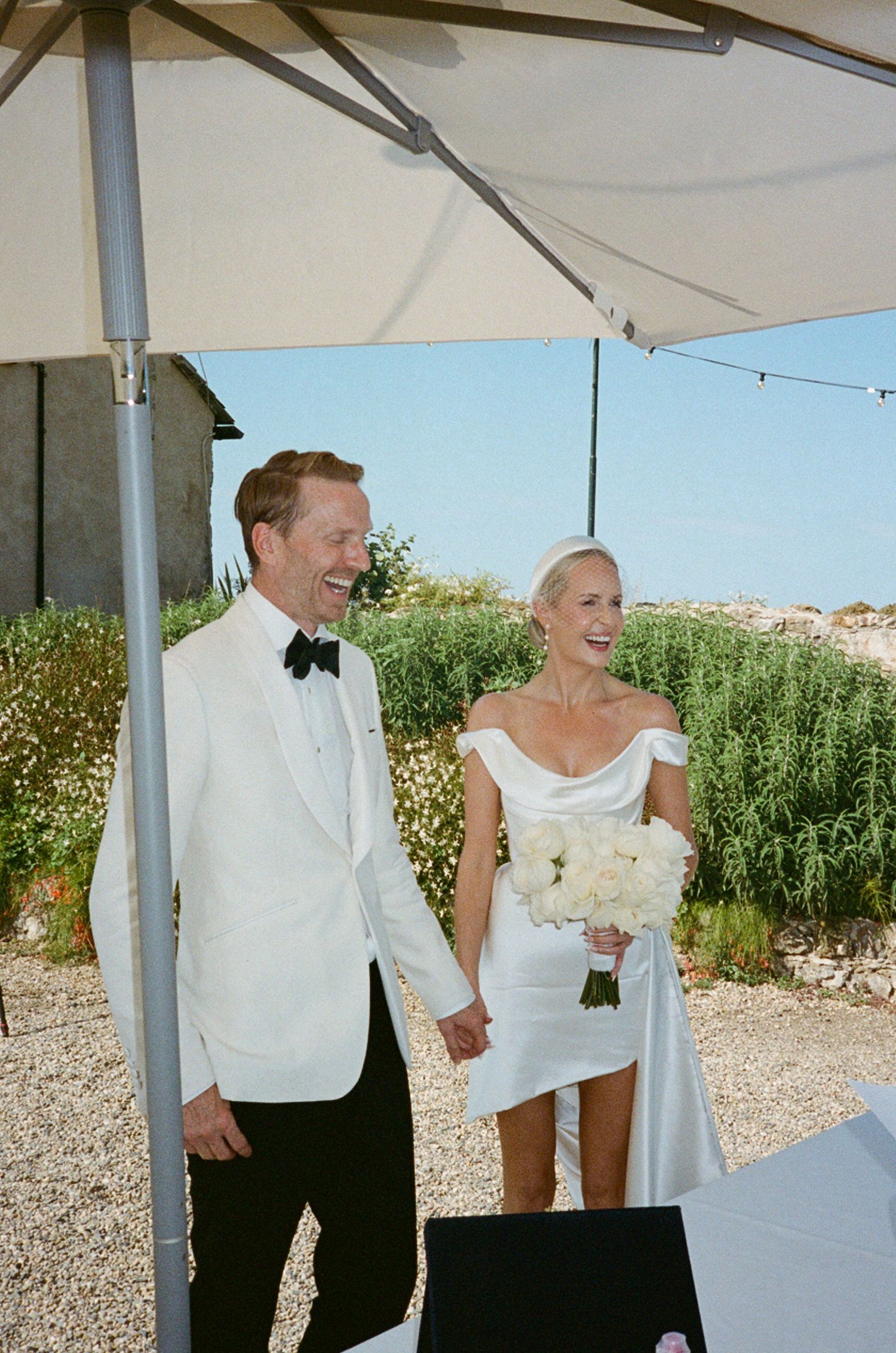 Happy couple in white wedding attire holding hands and smiling under outdoor umbrella.