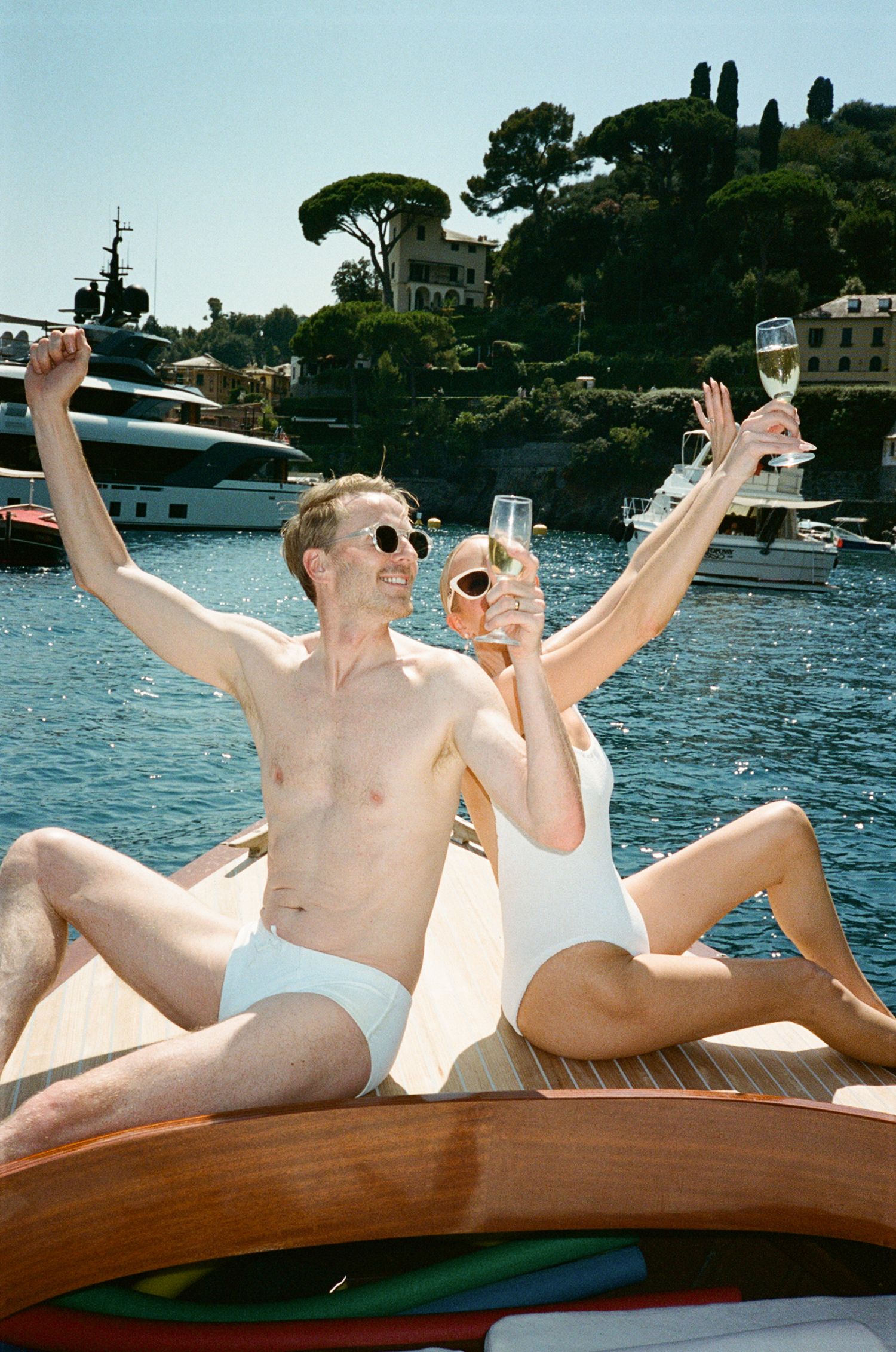 Joyful couple in white swimwear toasting champagne on a wooden boat in Portofino harbor, Italy.
