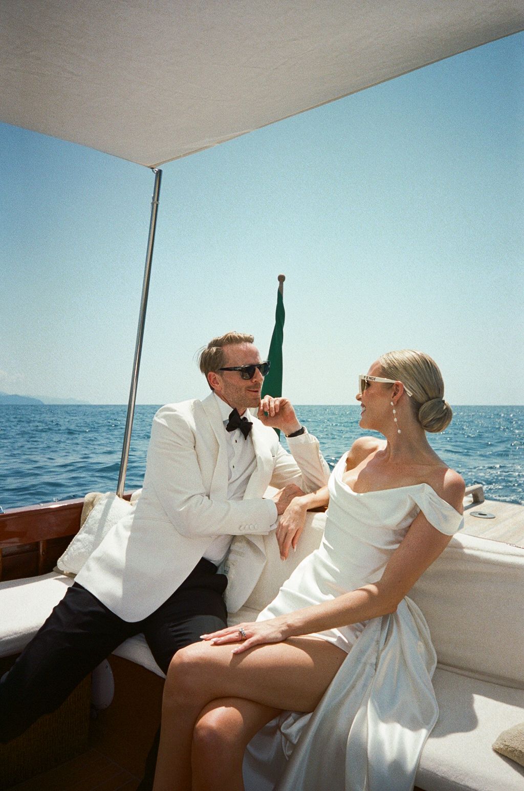 Stylish newlyweds in white wedding attire conversing on a wooden boat on the sparkling blue Mediterranean Sea.