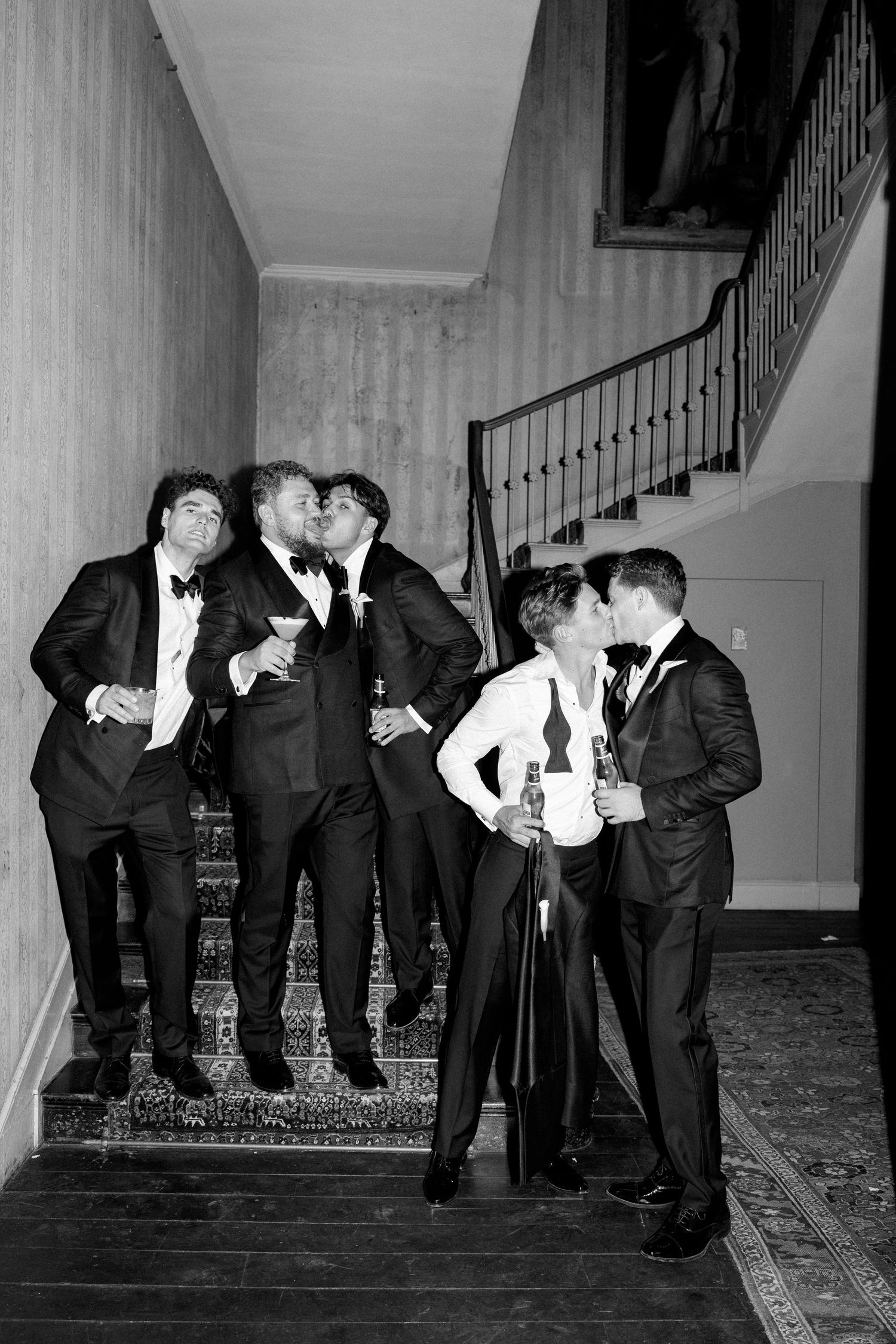 Black and white photo of groomsmen in tuxedos celebrating on a staircase, sharing kisses and holding drinks at a wedding.