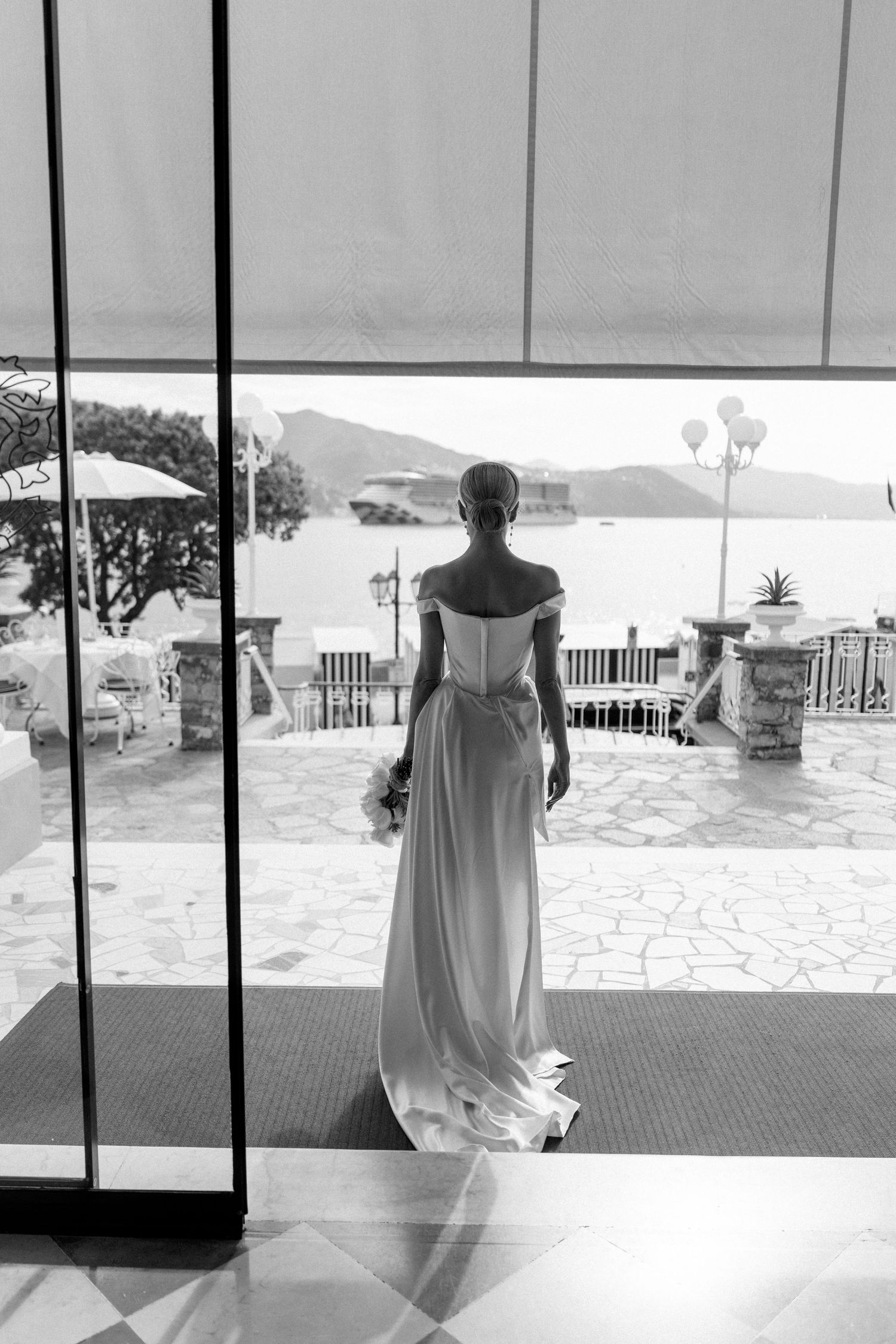 Black and white photo of a bride in an off-shoulder gown gazing at a Mediterranean waterfront through glass doors.