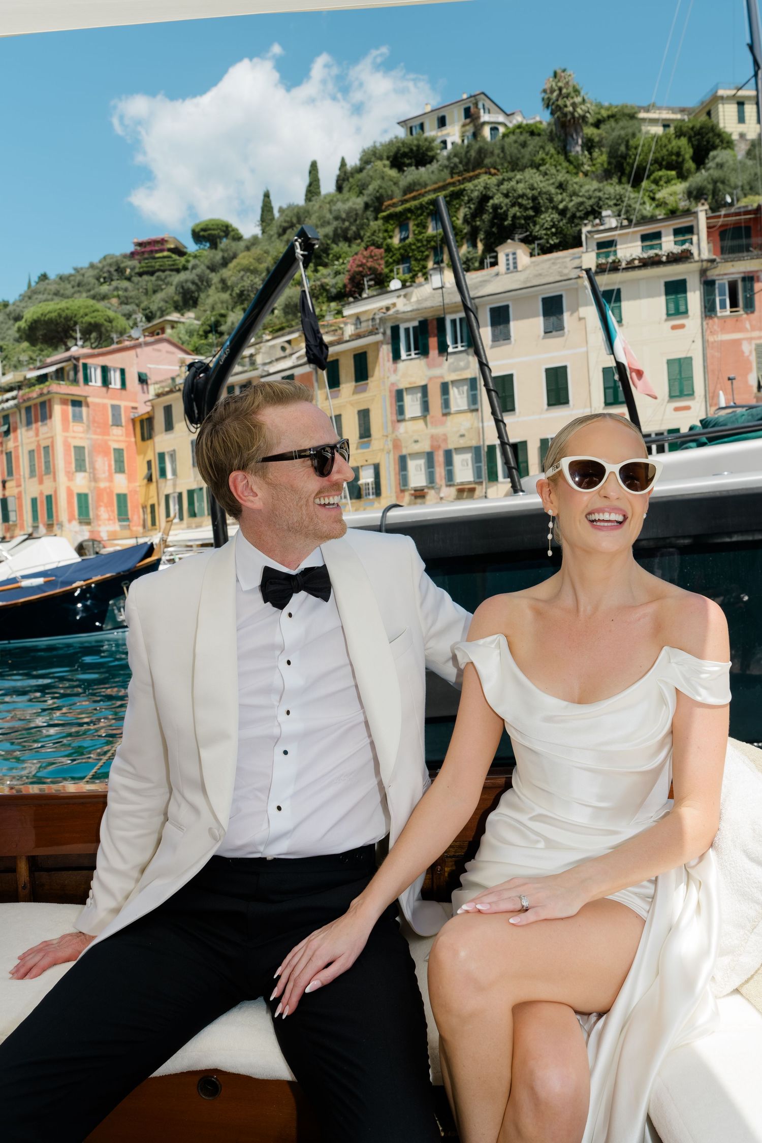 Smiling bride and groom in wedding attire sitting on a boat with Portofino harbor in background.