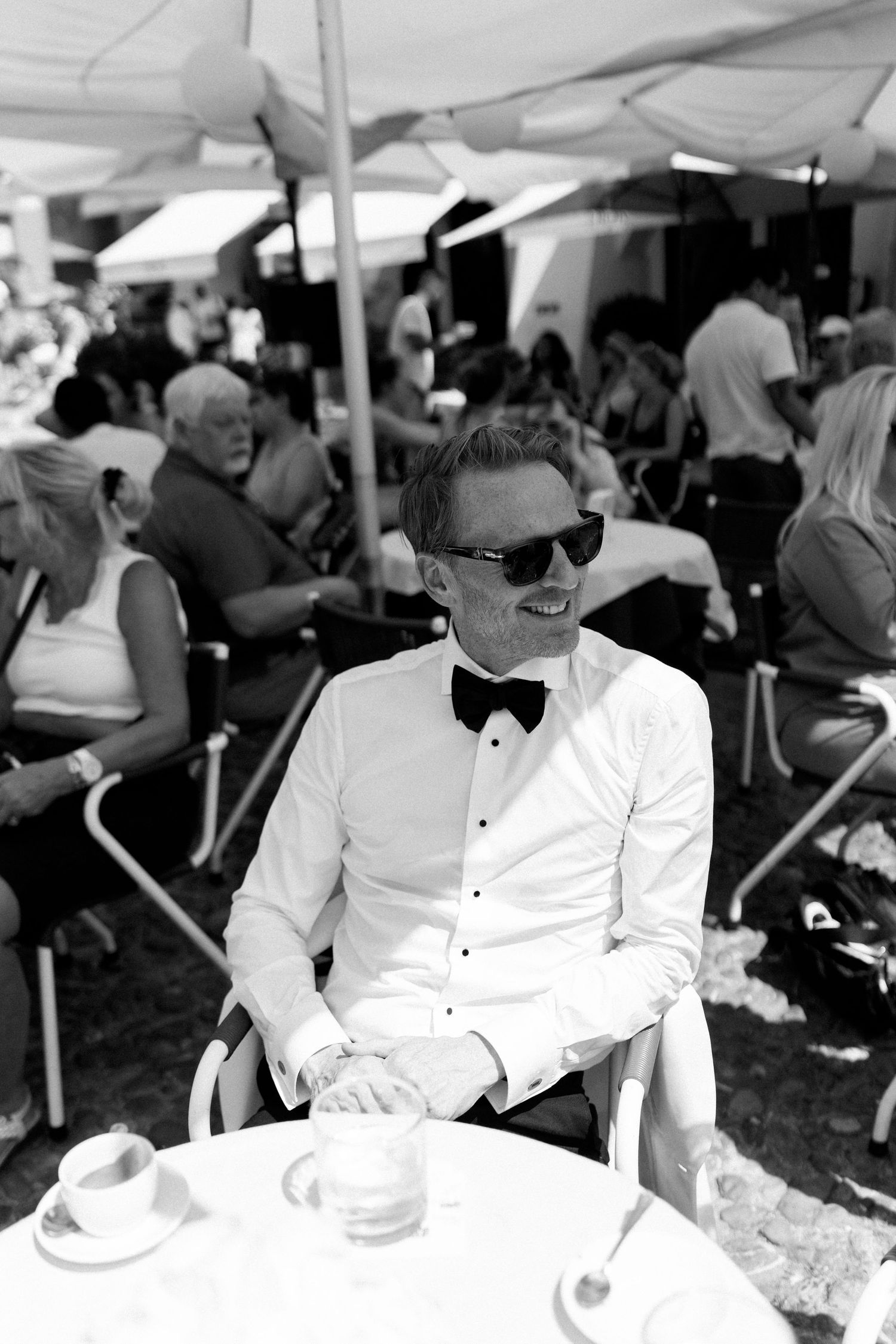 Elegant man in white tuxedo shirt and bow tie sits at outdoor cafe table wearing sunglasses.