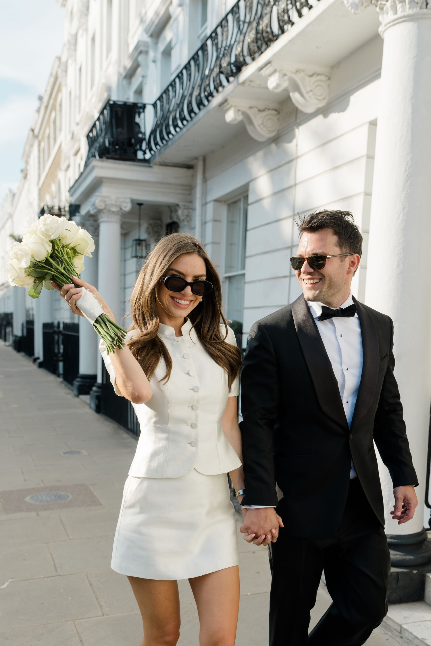 Stylish bride in white mini dress and groom in tuxedo walking hand in hand on elegant city street.