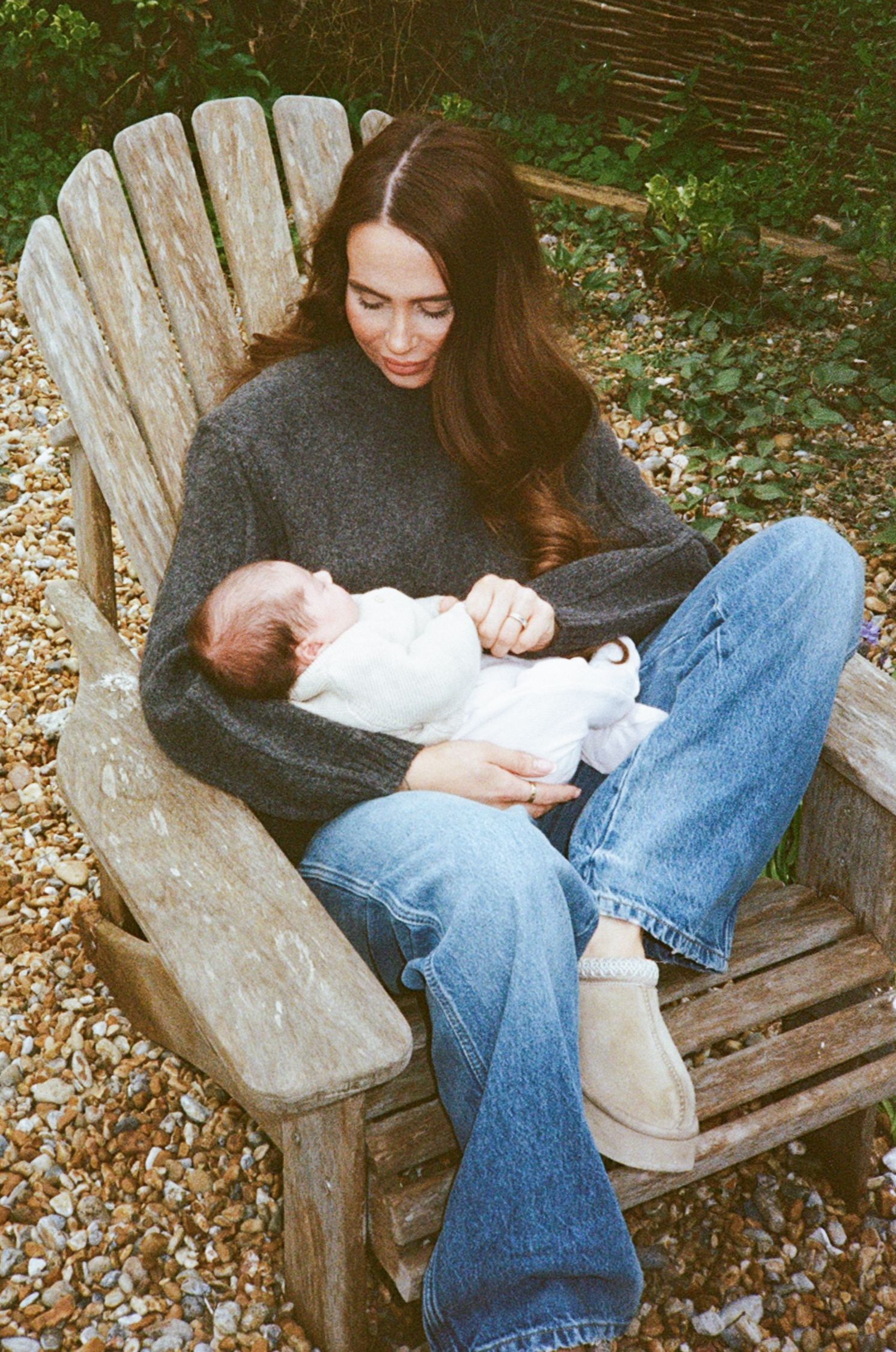 A young woman with long brown hair sits in a wooden Adirondack chair outdoors, cradling a sleeping newborn baby.