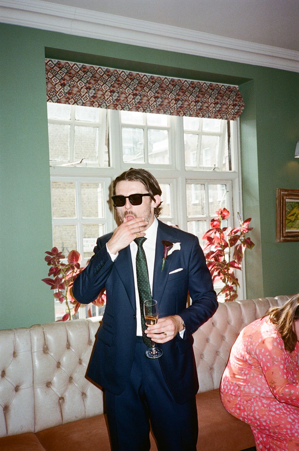 Man in navy suit and sunglasses holding champagne glass at an elegant indoor event with green walls.