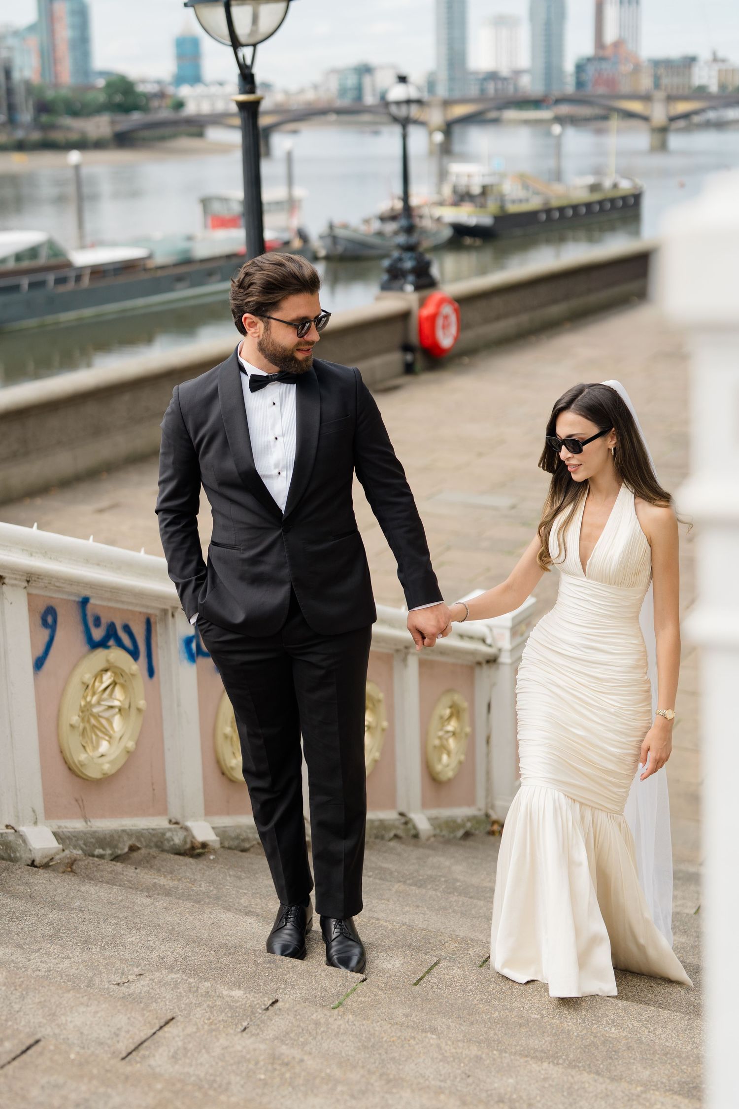 Elegant couple in formal attire holding hands on Thames riverside steps in London, man in tuxedo, woman in white gown.