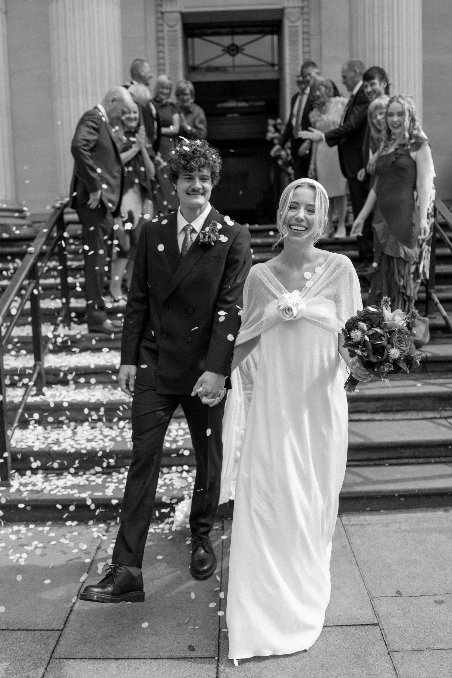Bride in flowing white dress smiles during confetti celebration outside historic building.