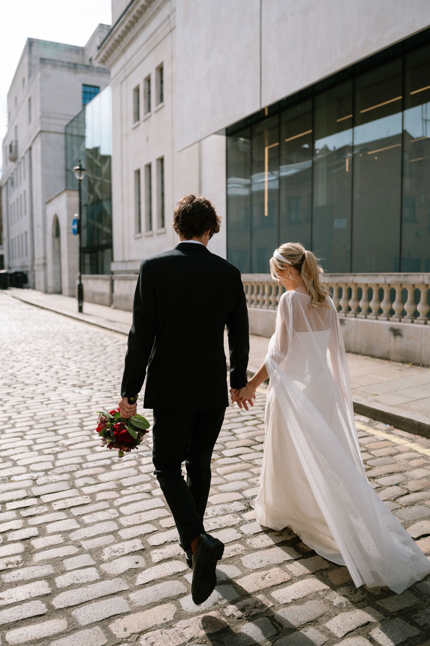 A bride and groom walk hand in hand on a cobblestone street with a bouquet of red flowers.