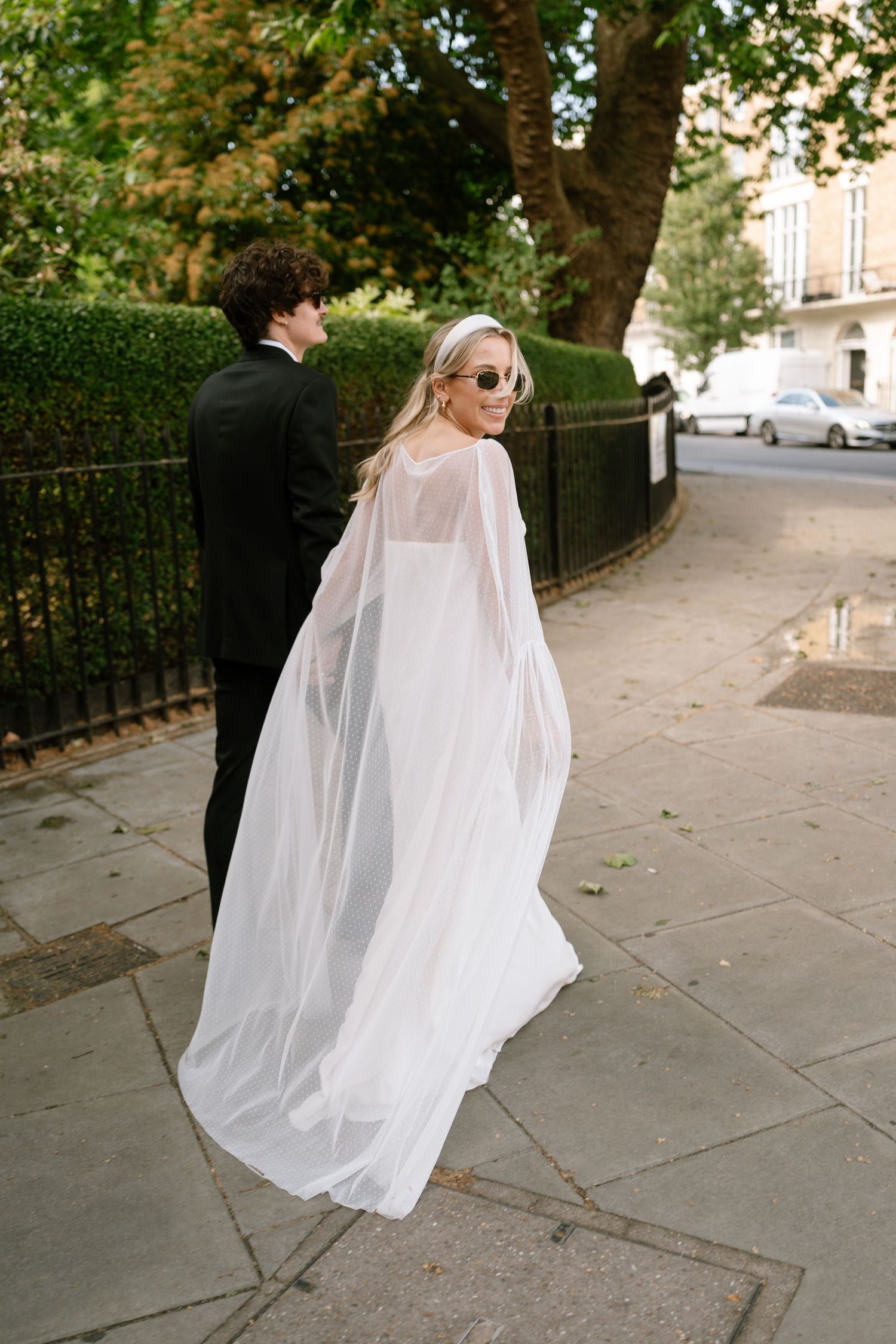 A flowing white dress catches the breeze on a London sidewalk lined with trees.