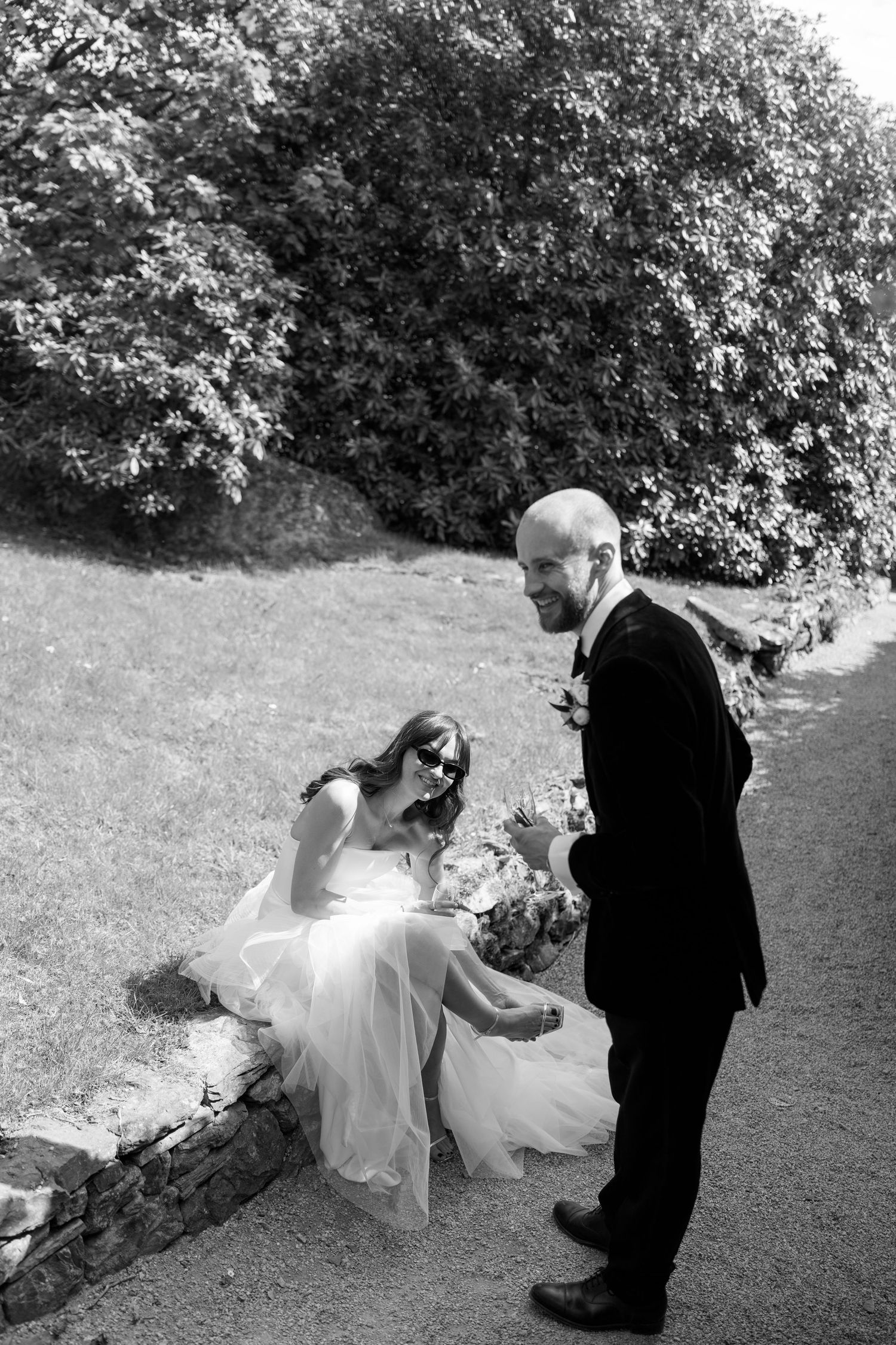 A couple in formal wedding attire poses playfully on a grassy lawn.