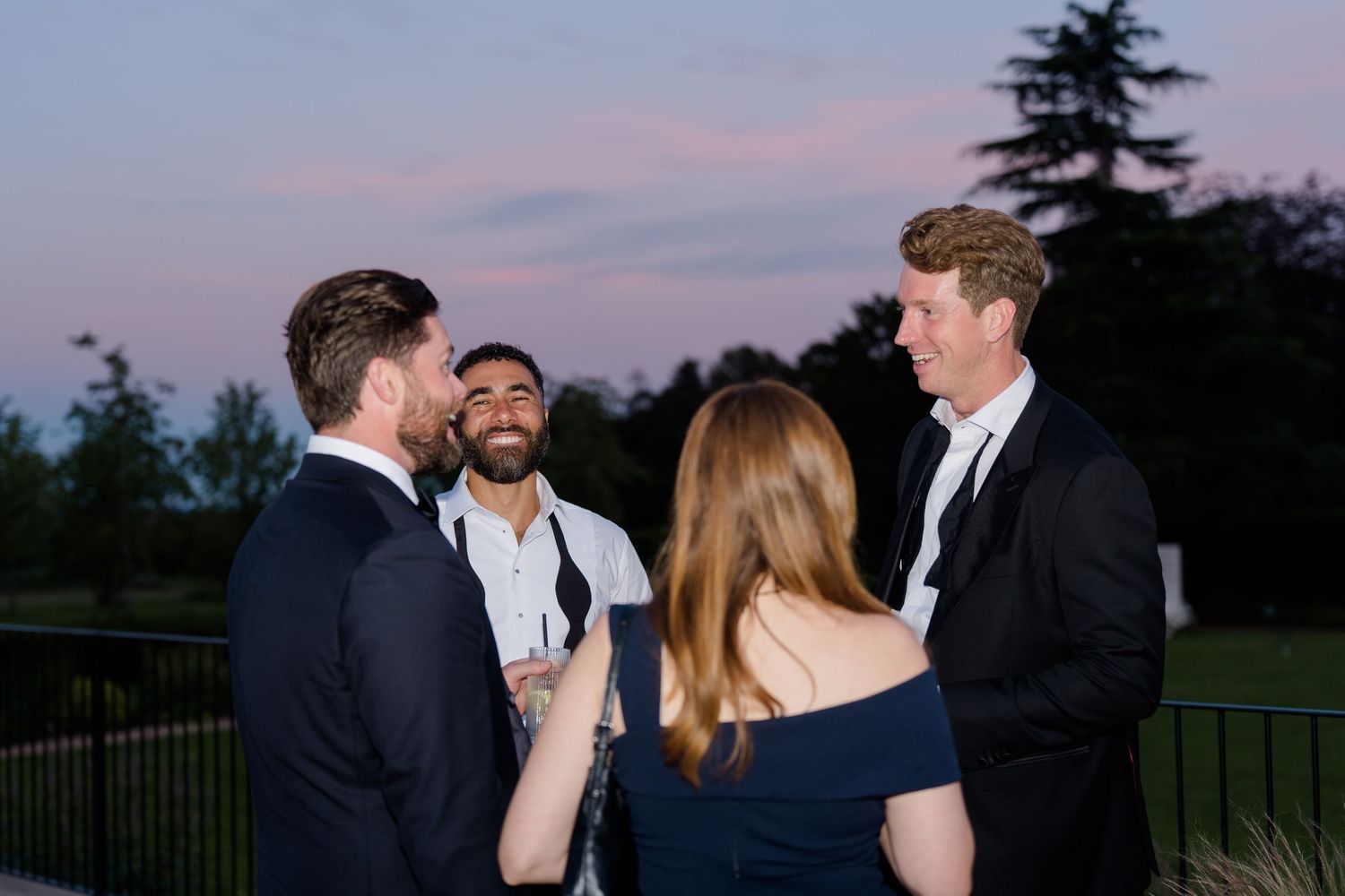 Group of elegantly dressed wedding guests laughing and conversing outdoors at dusk during evening reception.