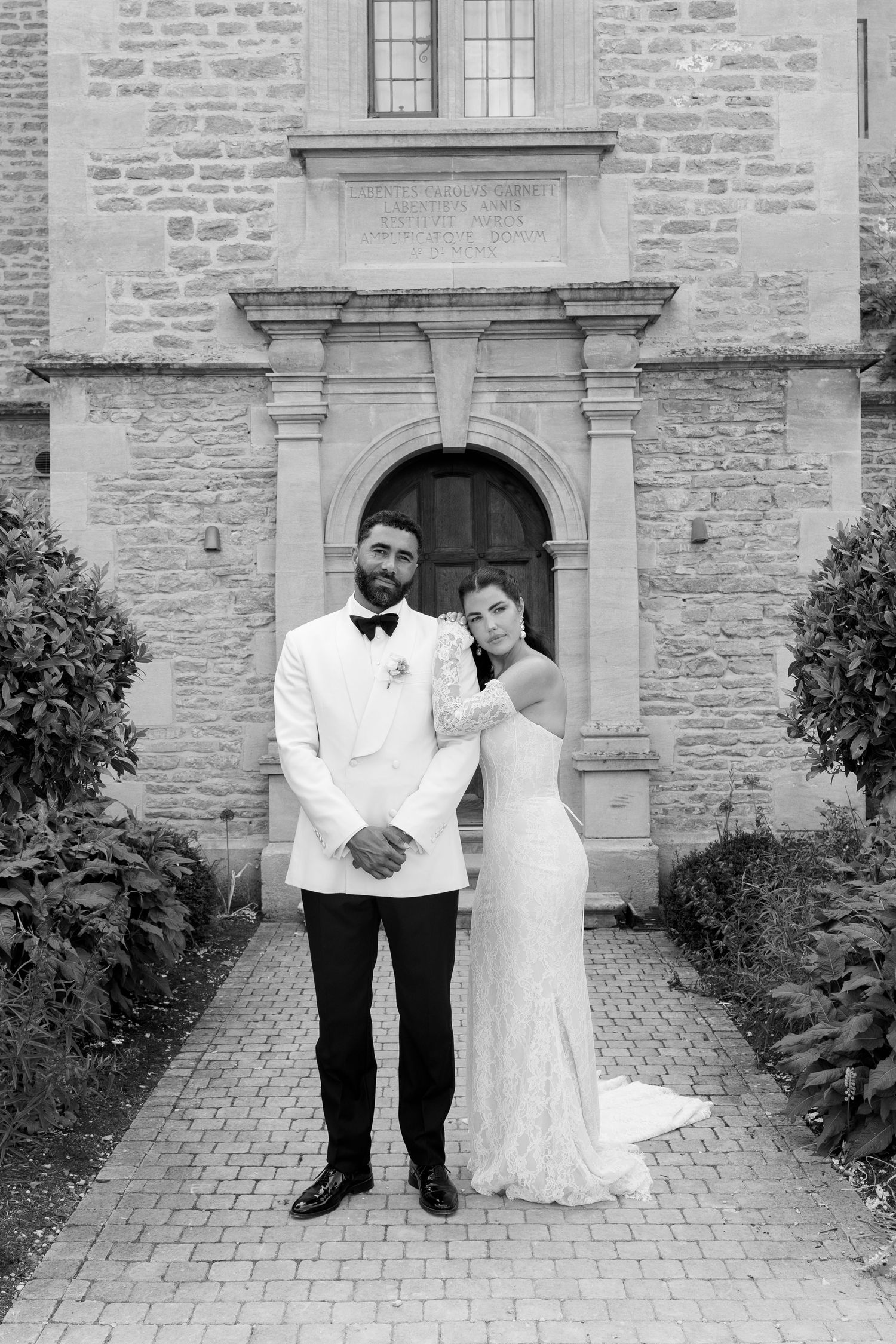 Bride and groom pose in black and white portrait before ornate stone church doorway.