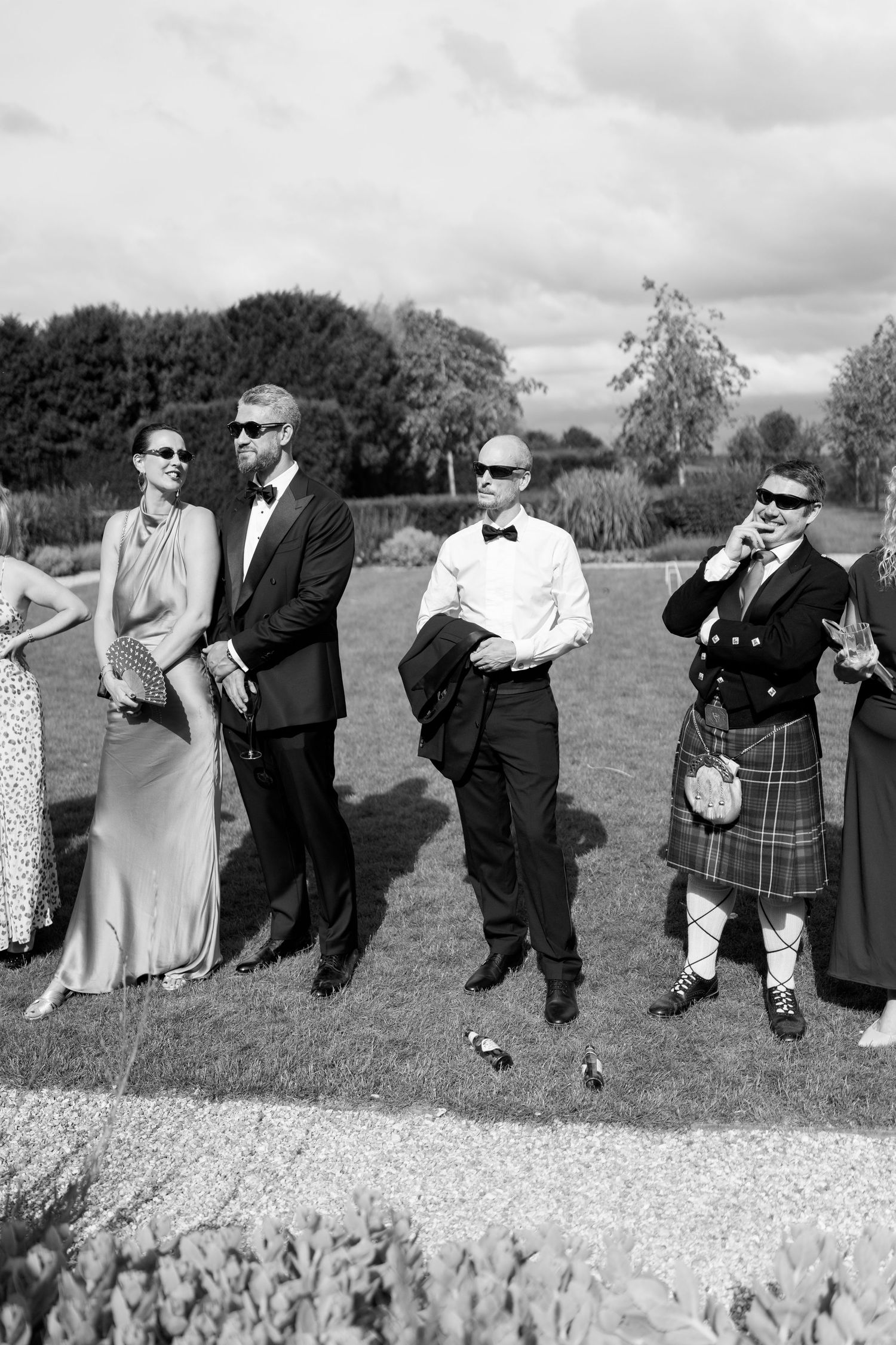 A group of elegantly dressed wedding guests including a man in a kilt pose outdoors in black and white.