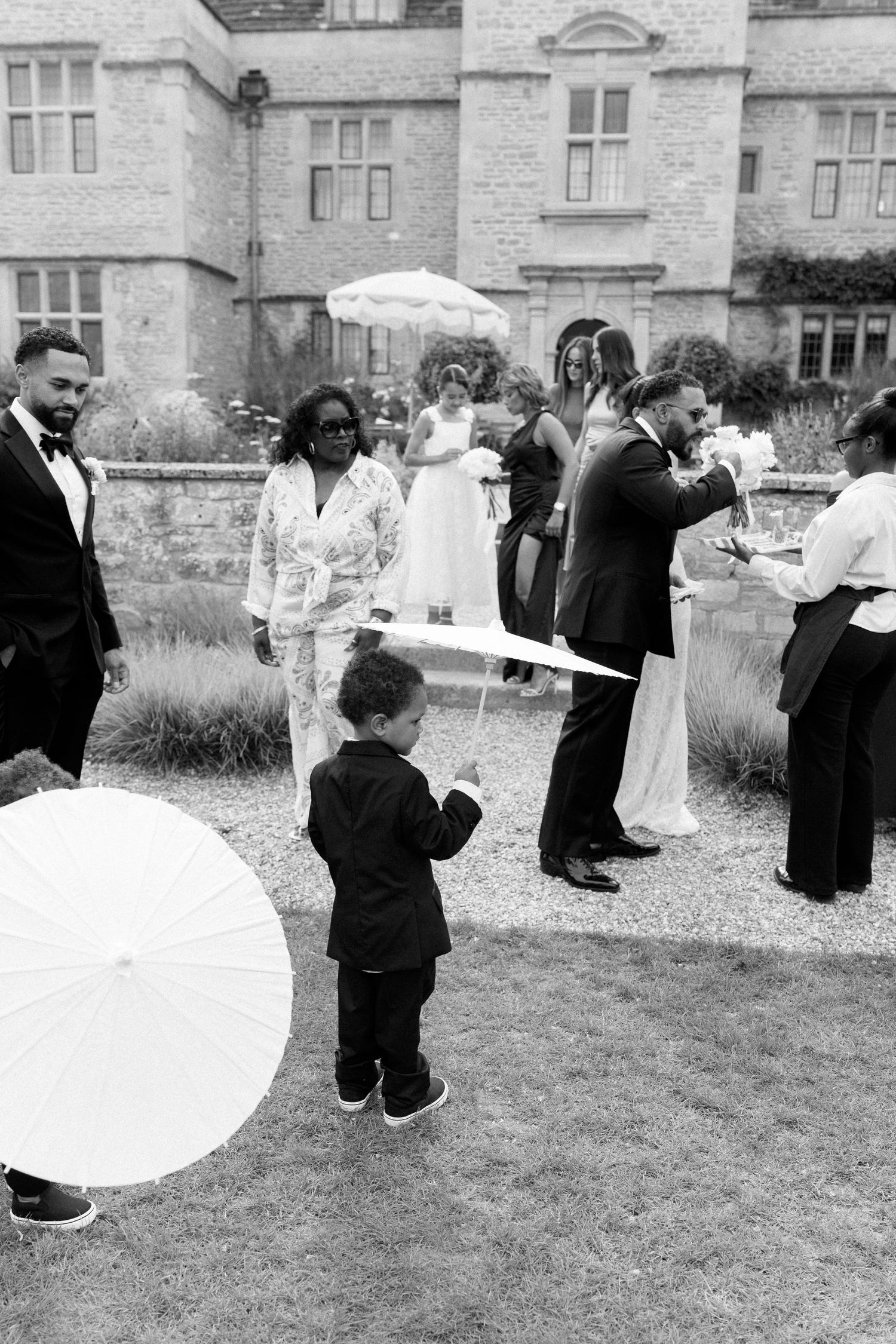 Black and white candid of wedding guests and child with parasols outside historic stone venue.